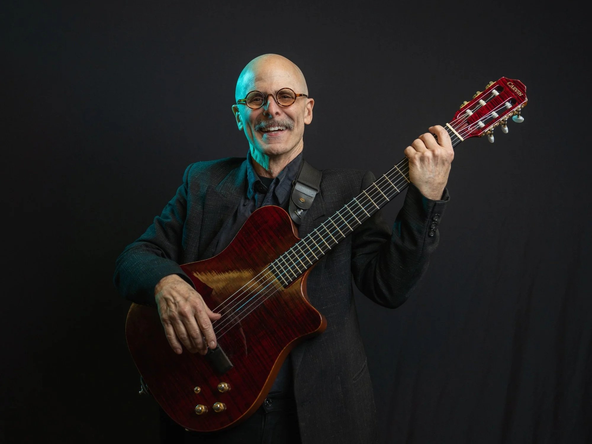 A smiling elderly man with glasses, wearing a black jacket, holding a red acoustic guitar against a dark background.