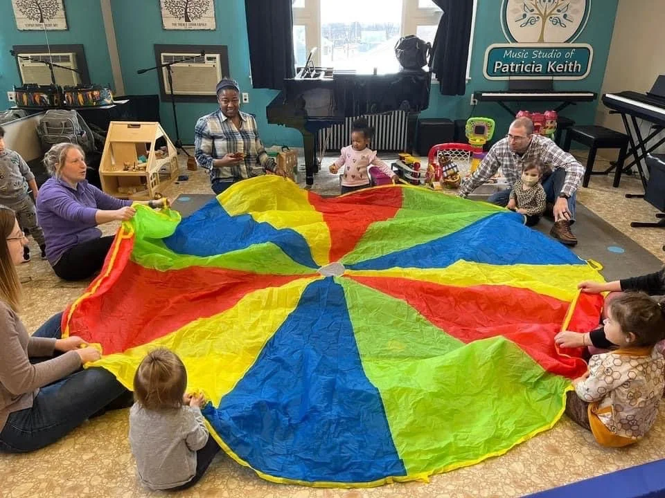 Ms. Jenni, the teacher, leads young students and their caregivers in a game using a colorful parachute during Busy Bees class.
