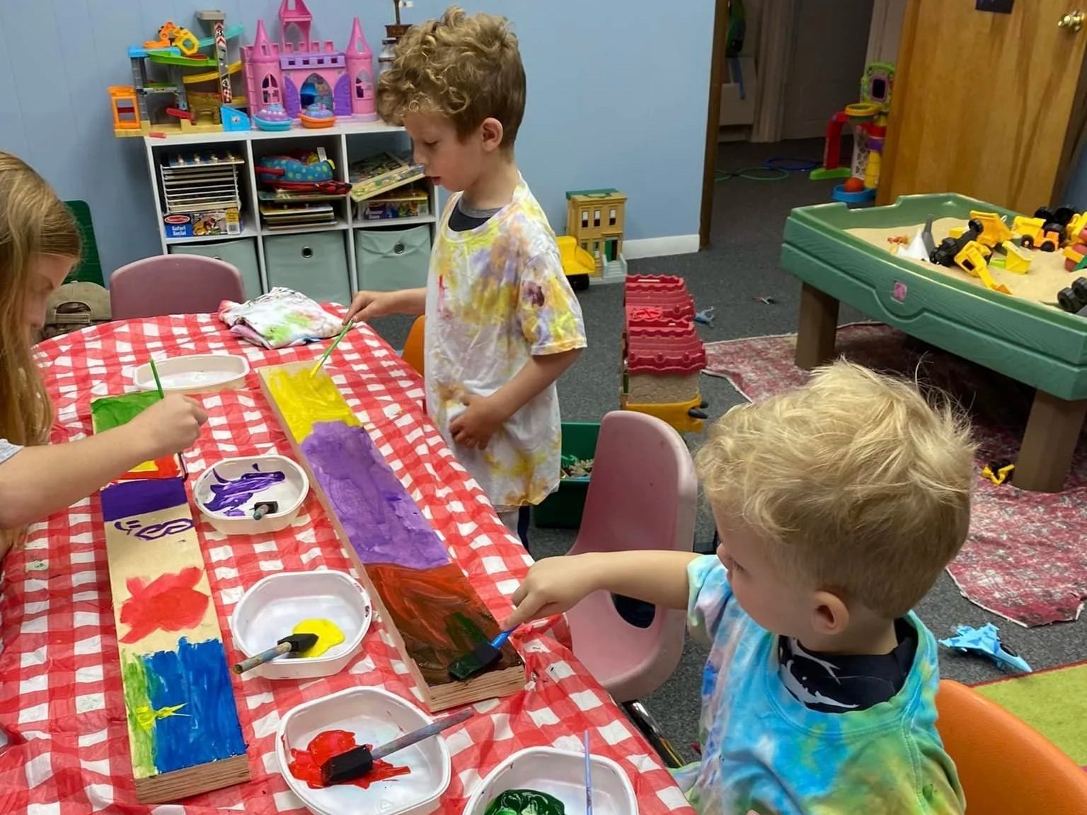 Children sit and stand at a table covered in a tablecloth, wearing smocks while they use brightly colored paint for an art project at our Boyertown studio during Art & Science Explorers class.