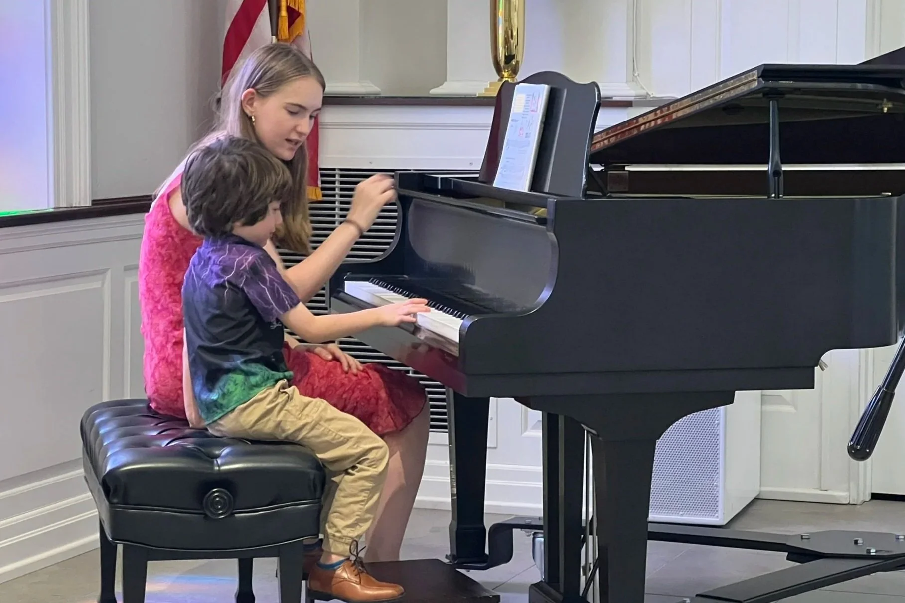 A teacher and student play a duet at a grand piano during a recital.