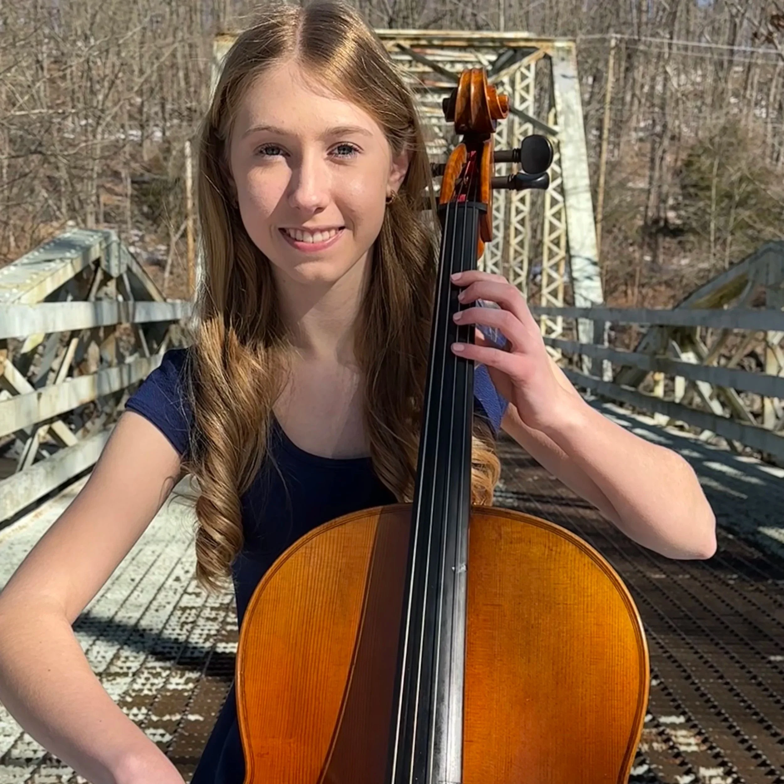 A young woman is outside holding her cello and bow while smiling