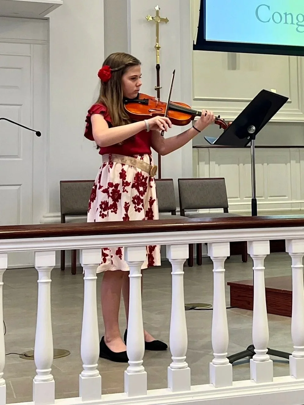 A girl playing violin in a formal setting, wearing a red top, floral skirt, black shoes, with a red flower in her hair.