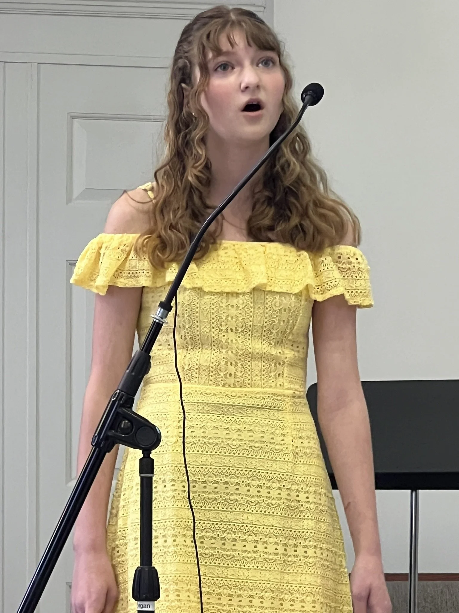 Young woman with curly blonde hair singing into a microphone, wearing a yellow off-shoulder dress.