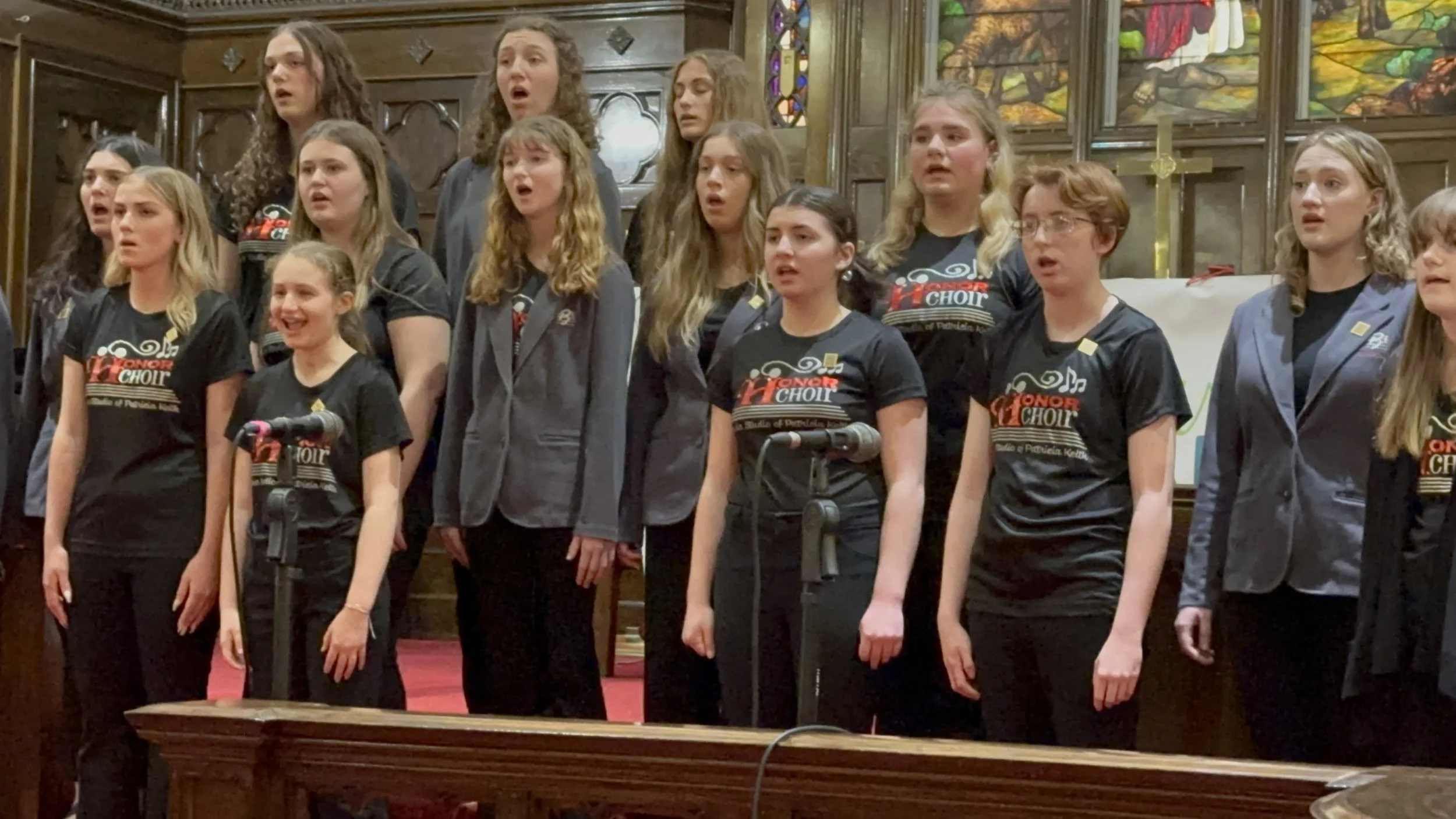 Students singing in choir, standing behind microphones in a church with stained glass windows.