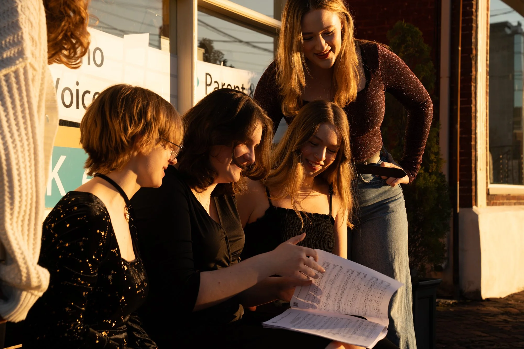 Five young women gathered outdoors around their sheet music, smiling and looking at it together during golden hour sunlight.