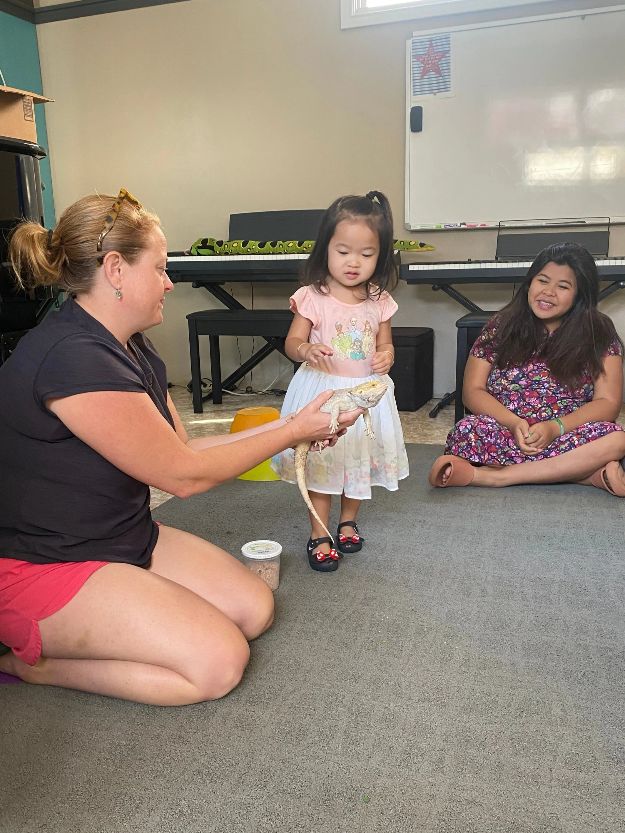 A photo of a woman kneeling on the floor, holding an off-white lizard for a toddler to touch
