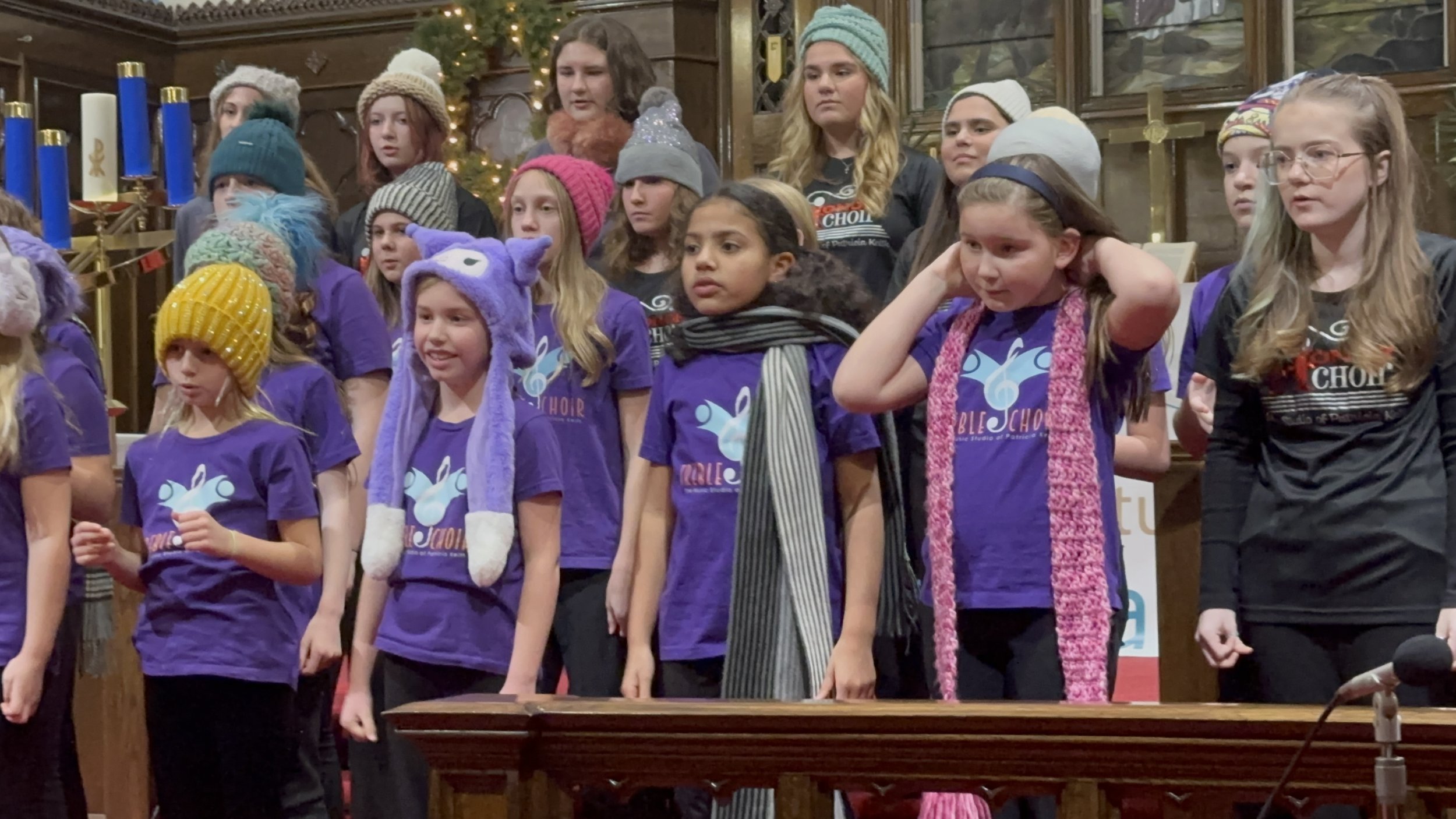 A group of young singers in the Treble Choir ensemble wearing winter hats & scarves is performing.