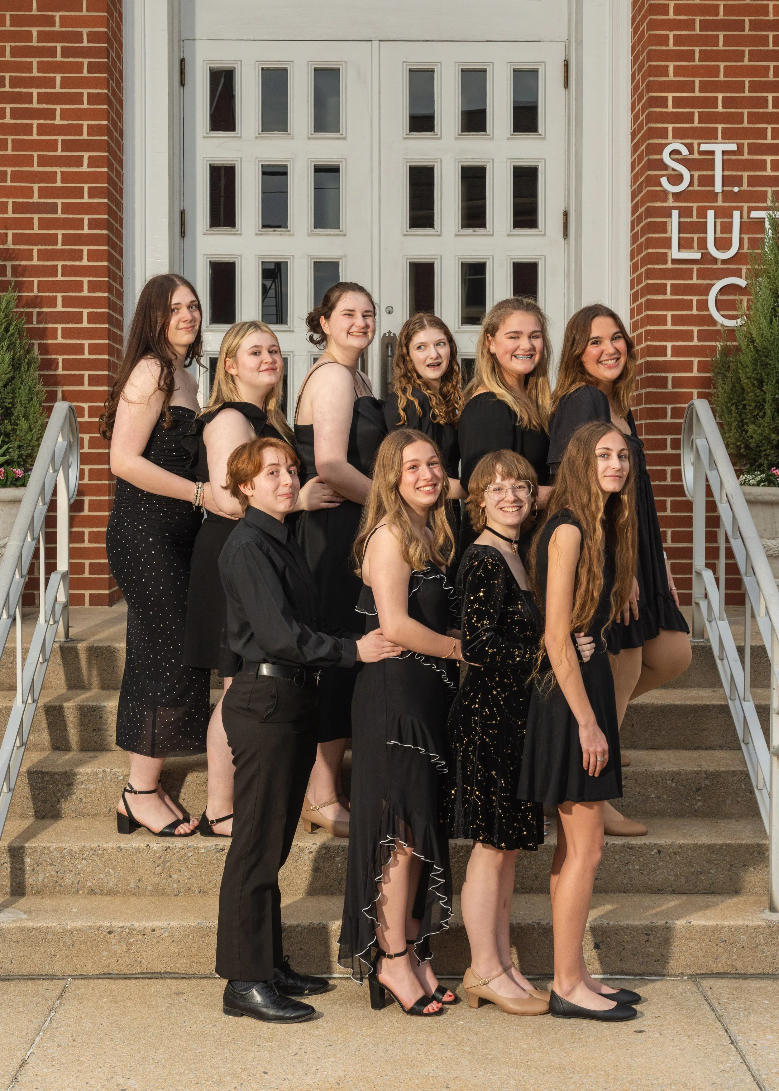 Group of eleven young women standing on steps outside a brick building, dressed in black formal attire, smiling for a photo.