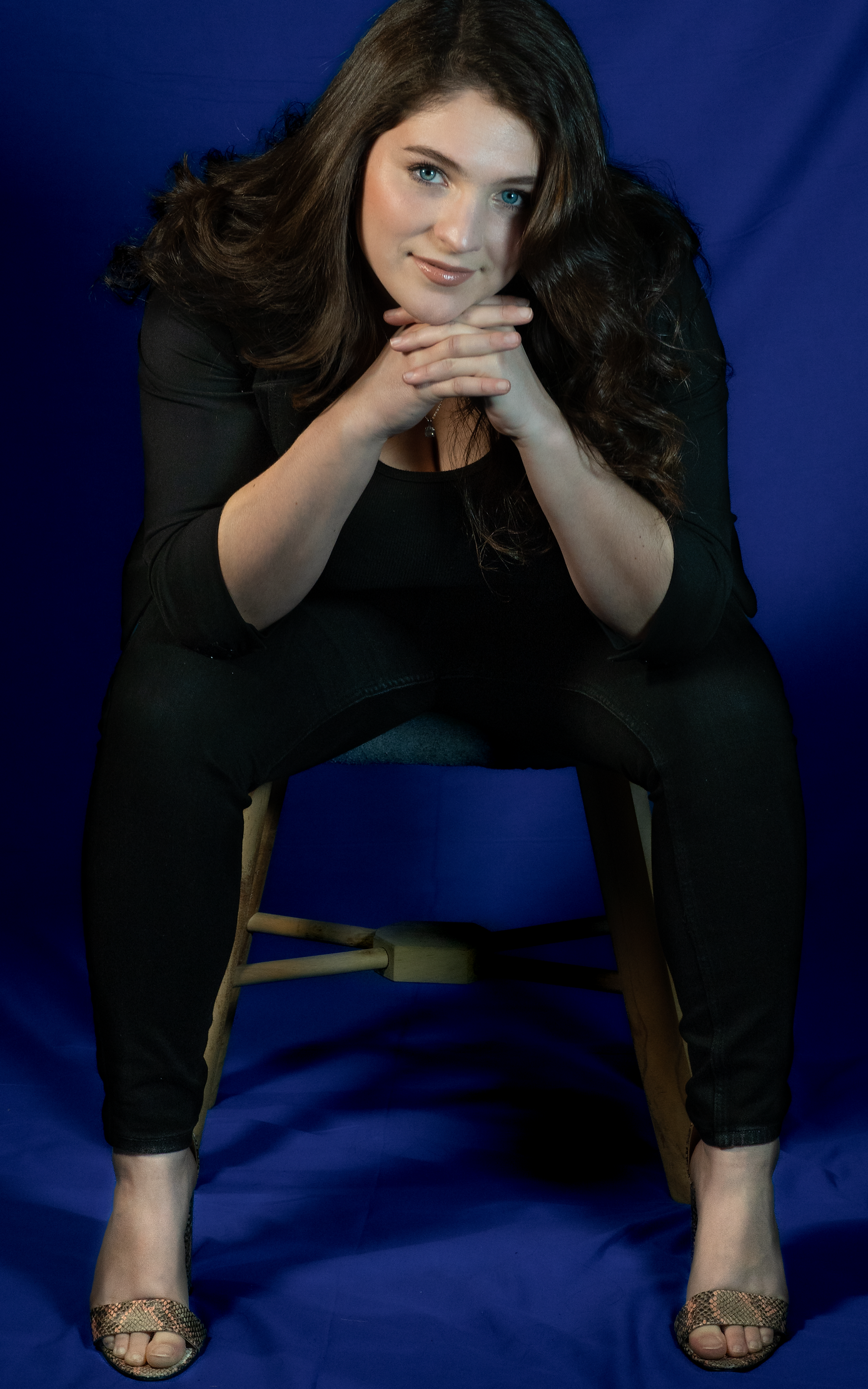 A full body shot of a seated woman with dark hair, smiling with her hands clasped under her chin