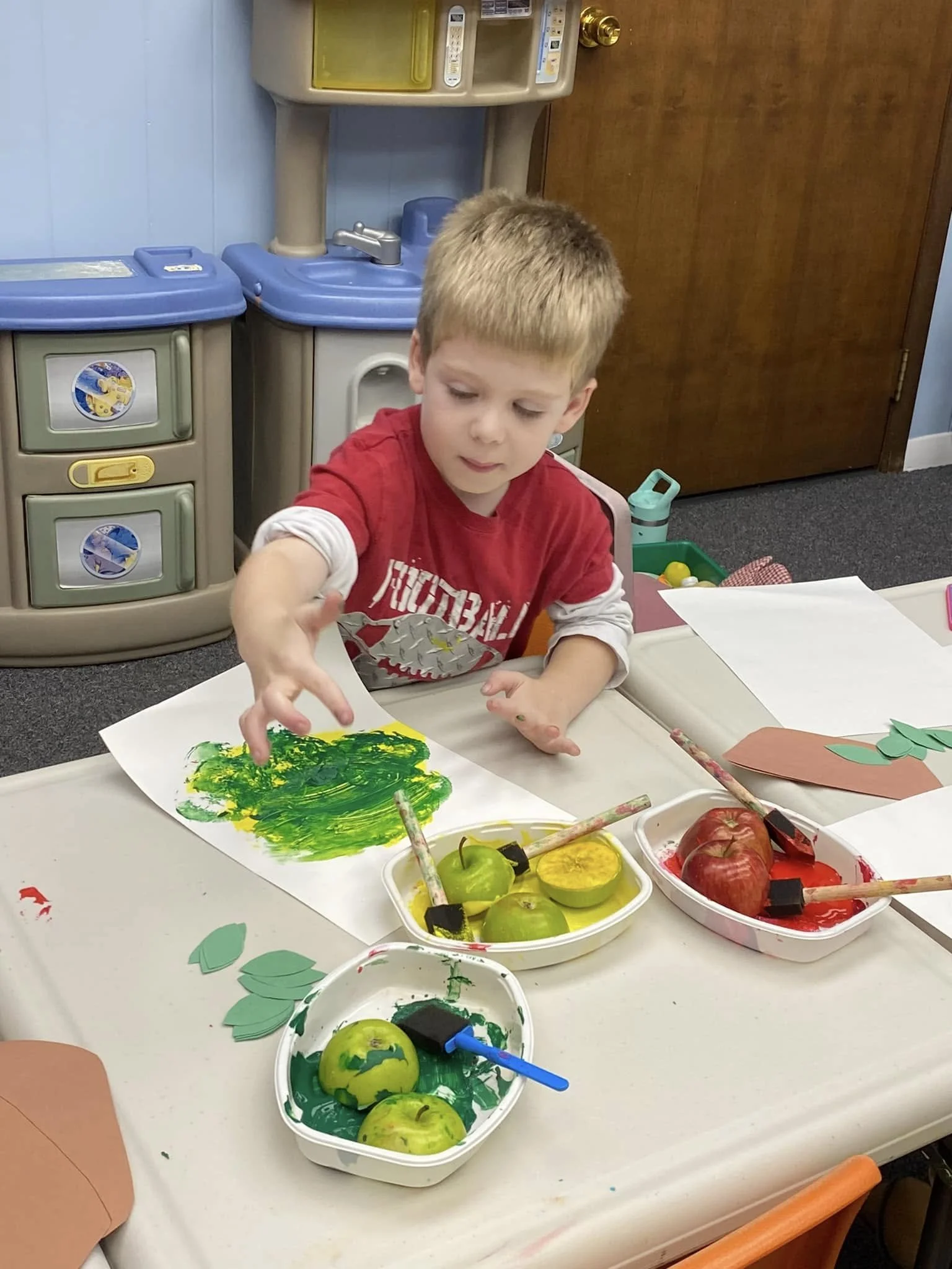 A young child uses apple slices and green and yellow paint for his artwork during class.