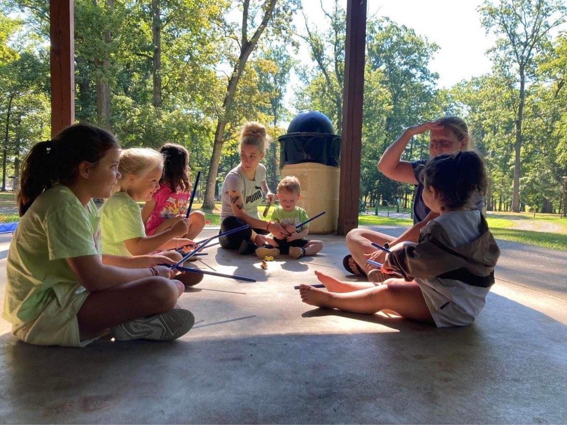 Children with teacher seated in a circle outside under a pavilion