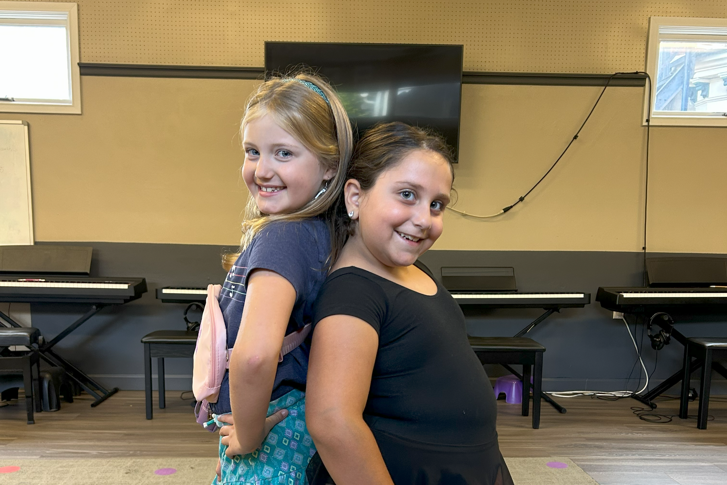 Two young girls smiling and posing back-to-back in a room with pianos and a large screen.