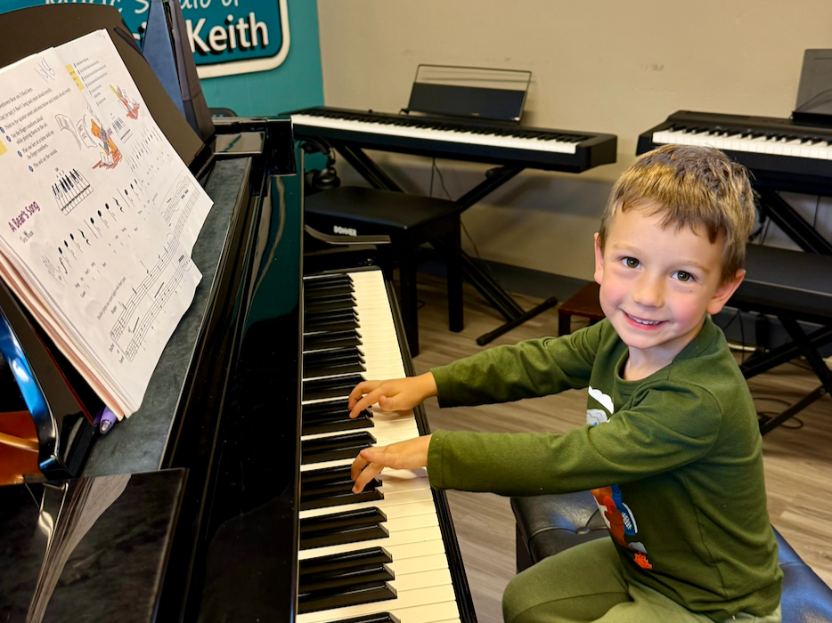 A smiling boy with light hair sits at a piano with his hands on the keys.