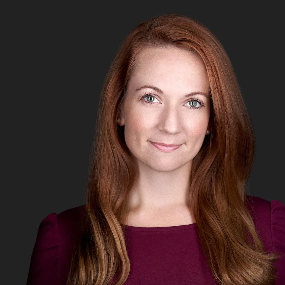 A professional photo of a smiling woman with red hair against a plain dark background
