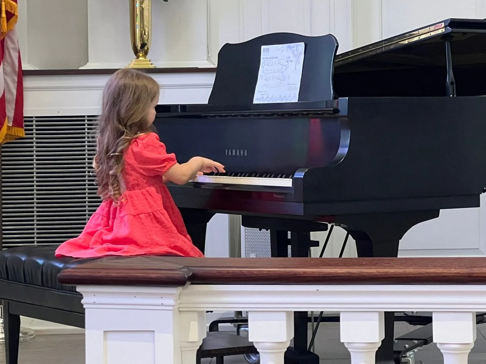 A young girl in a pink dress sits at a black grand piano.