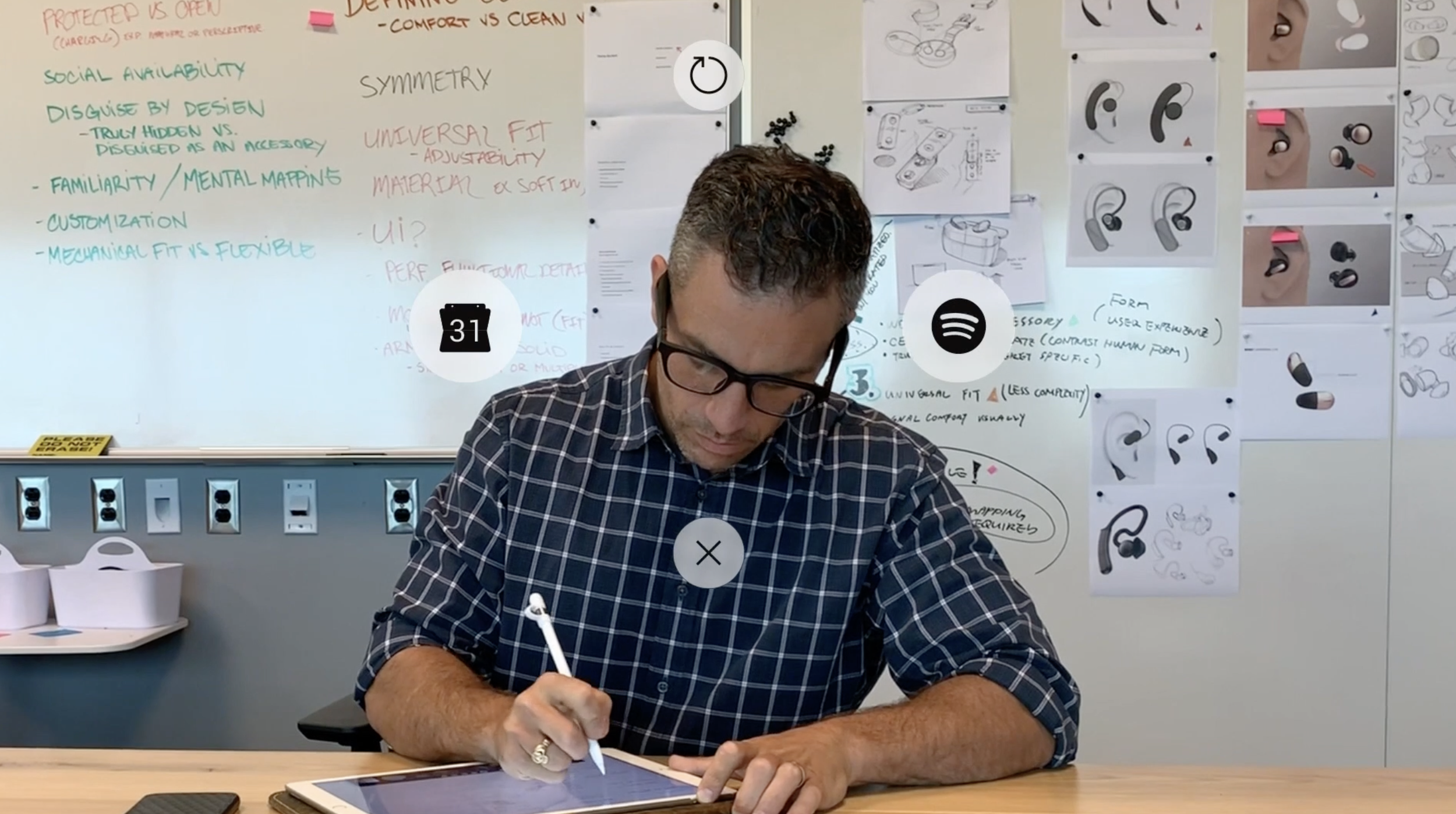 A man in glasses and a plaid shirt is sitting at a table writing on a tablet with a stylus. Behind him, a whiteboard with colorful handwritten notes and diagrams related to design and user interface is visible, along with sketches of earbuds and headphones taped to the wall.
