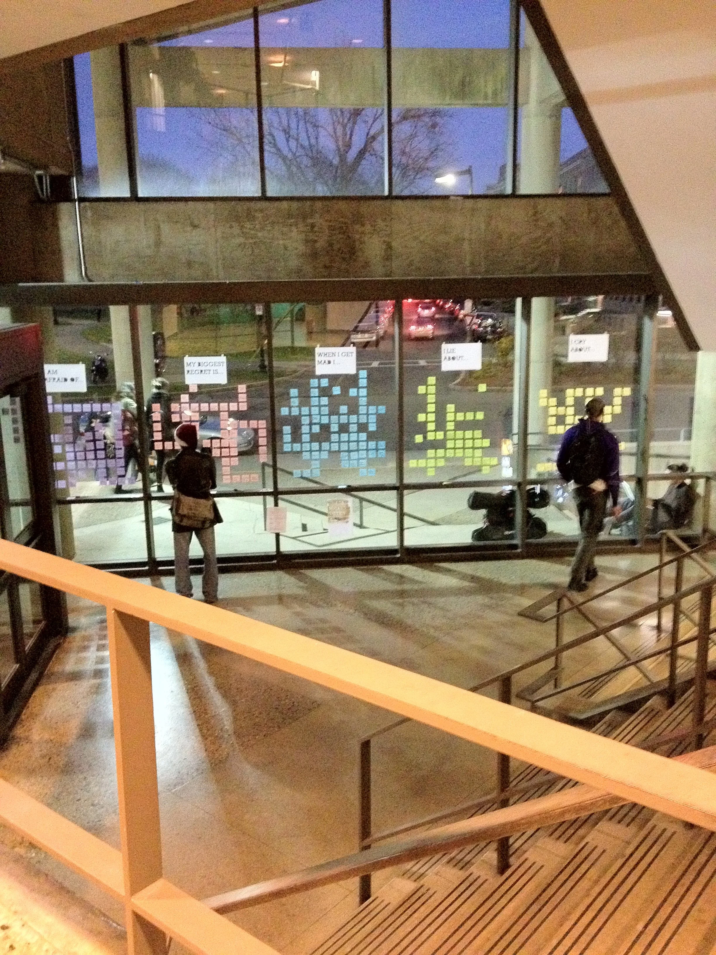 A view of a modern building's large glass window with colorful sticky notes arranged to form pixelated words or phrases. Inside, people are looking at the notes, and outside, a tree and cars can be seen through the window during dusk or evening.