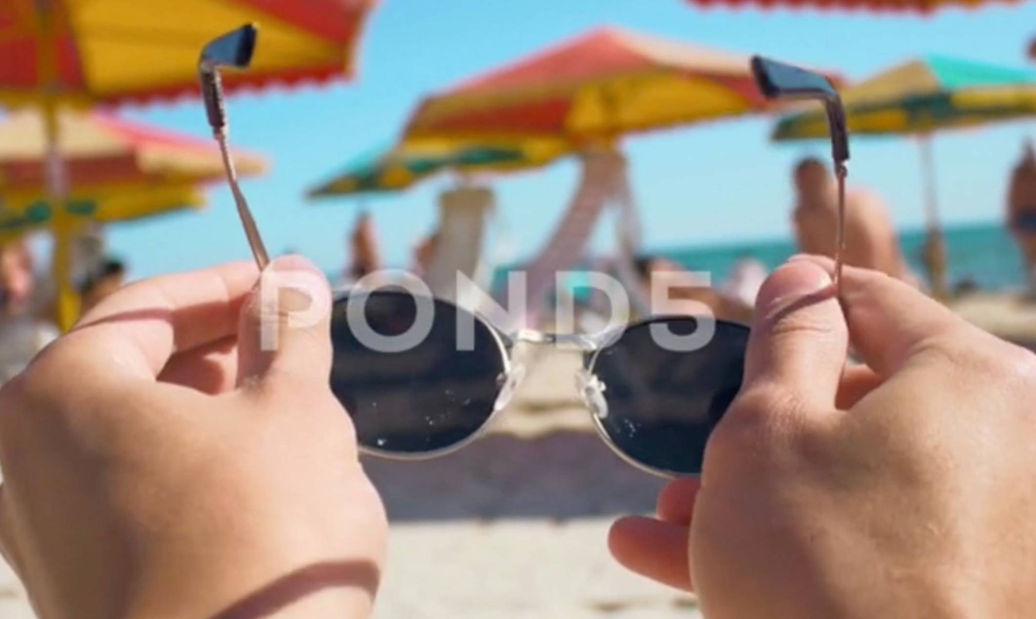 Close-up of hands holding sunglasses on a beach with colorful umbrellas in the background.