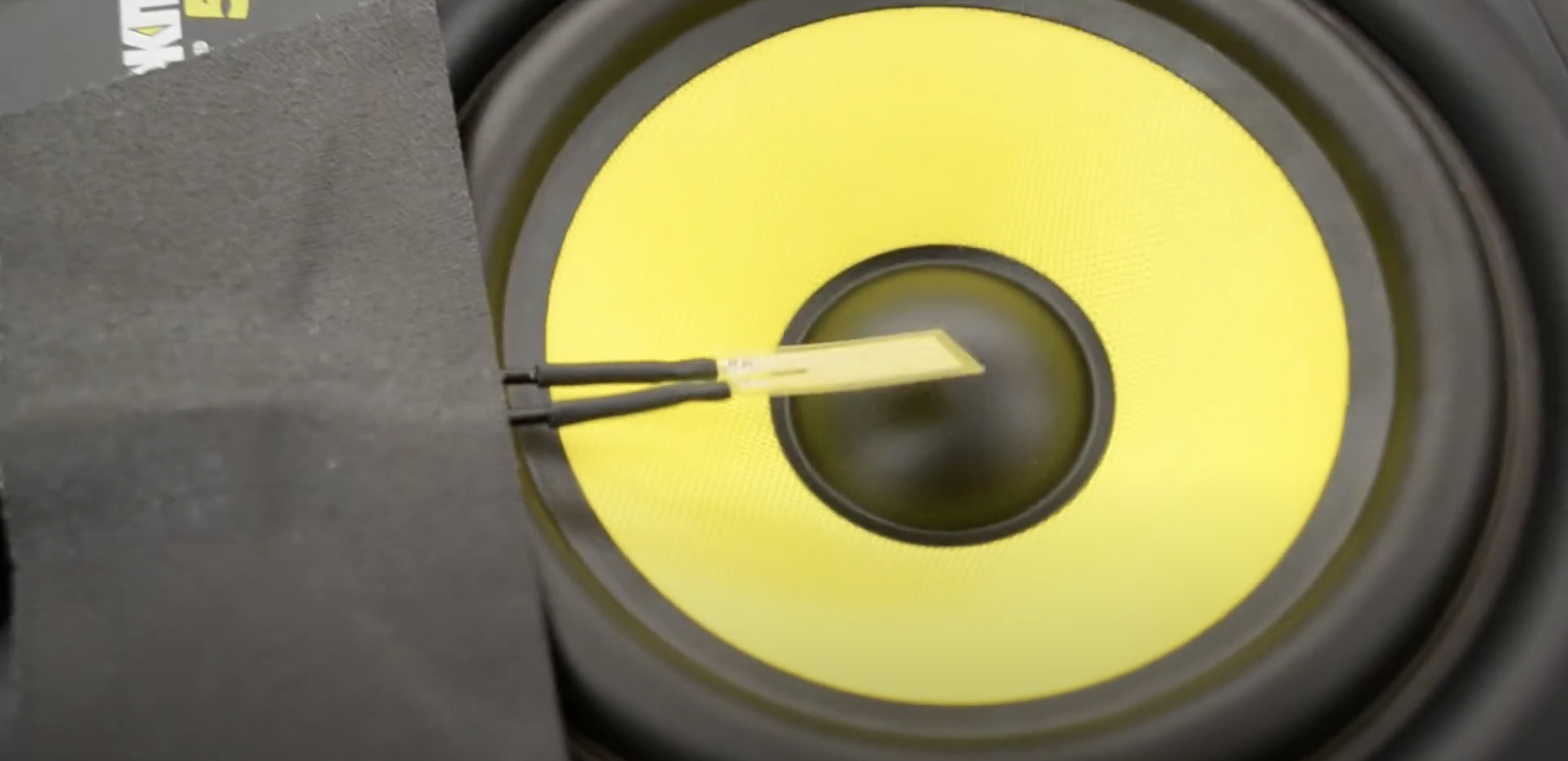 Close-up of a yellow speaker with two black wires and a small yellow battery clip on top.