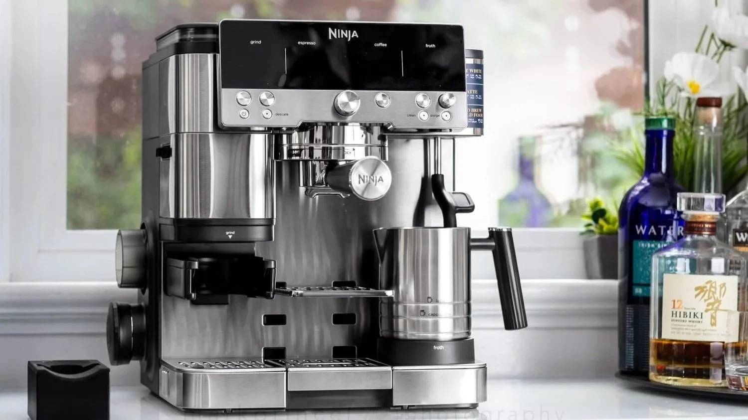 A stainless steel Ninja coffee machine and espresso maker placed on a kitchen countertop with bottles, flowers, and a window in the background.