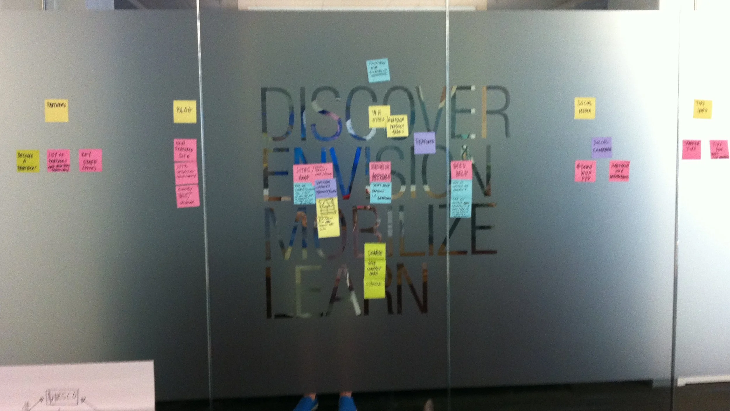 A gray wall with the words 'DISCOVER EVOLVE INSPIRE' cut out of the metal surface. Pink and yellow sticky notes are arranged around the words with various handwritten notes.