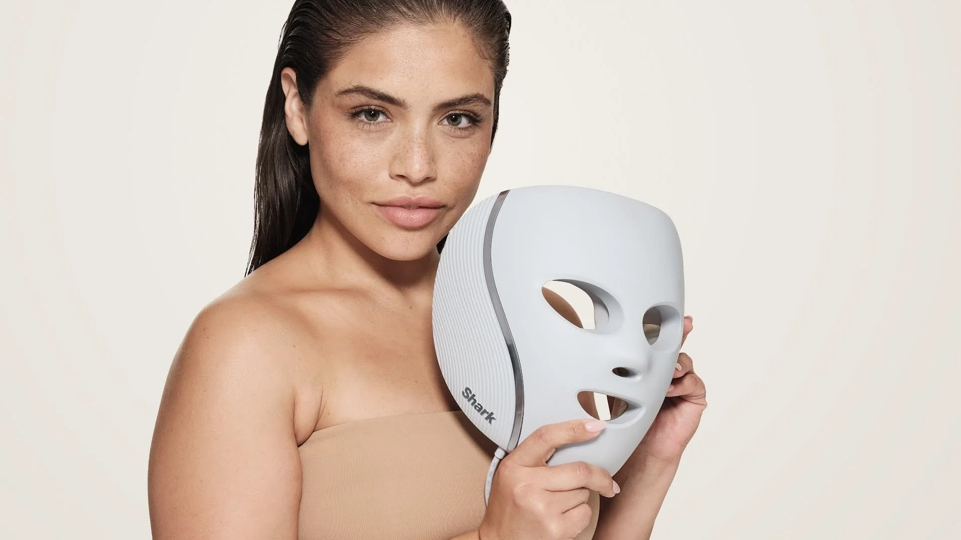 A woman with dark brown hair and clear skin holding a white facial mask with eye and mouth openings, against a plain light background.