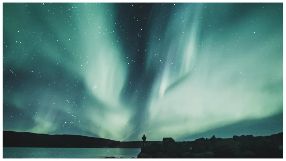 Night sky with vibrant green Northern Lights over water and a person standing on the rocky shoreline.