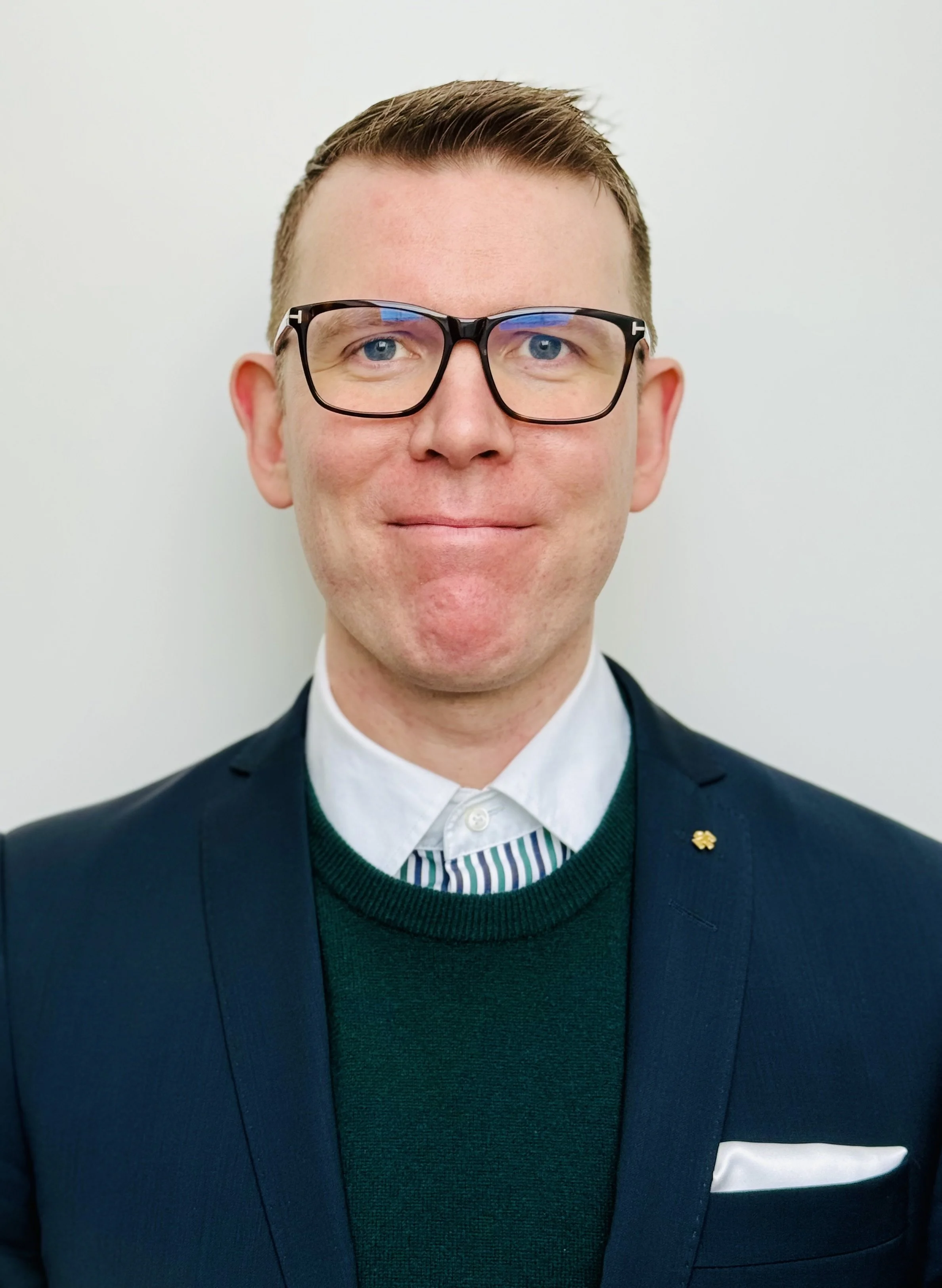 A man with glasses wearing a dark suit jacket, white shirt, and a green sweater, standing against a plain white background.
