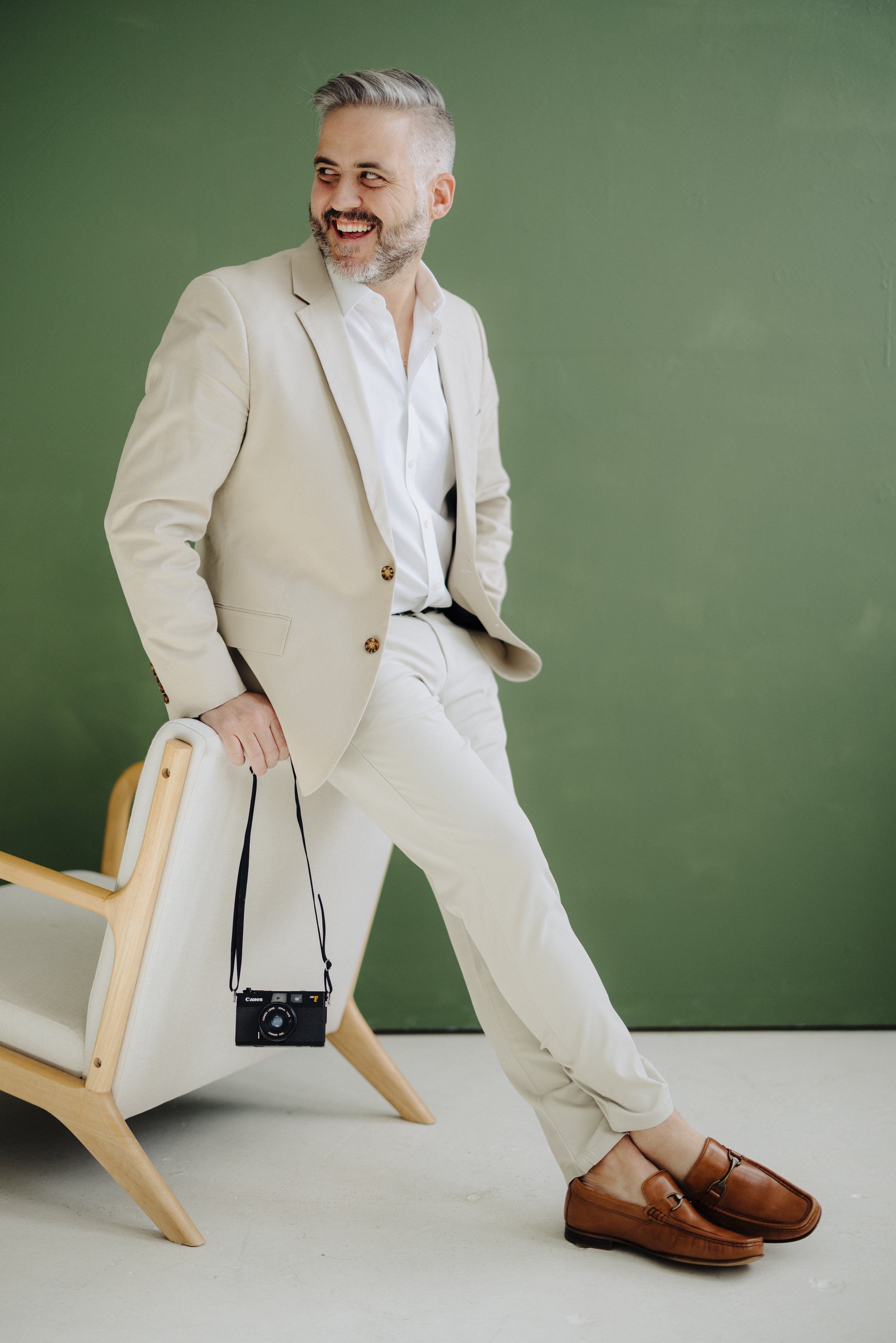 A man with gray hair and beard wearing a cream-colored suit and white shirt sitting on a chair with a Canon camera hanging around his neck, in front of a green wall.