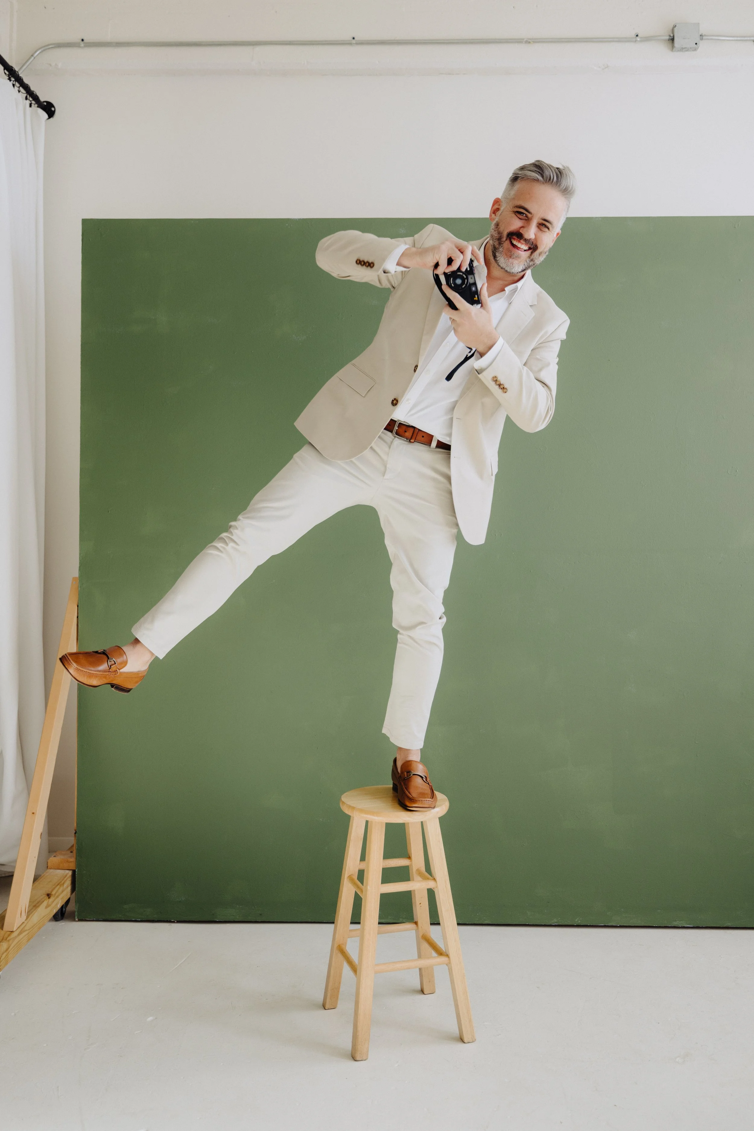 A man in a white suit standing on a stool, balancing with one foot on the stool and the other extended, holding a camera towards the camera taking his photo. He is smiling against a green background, with a white wall and curtain on the side.