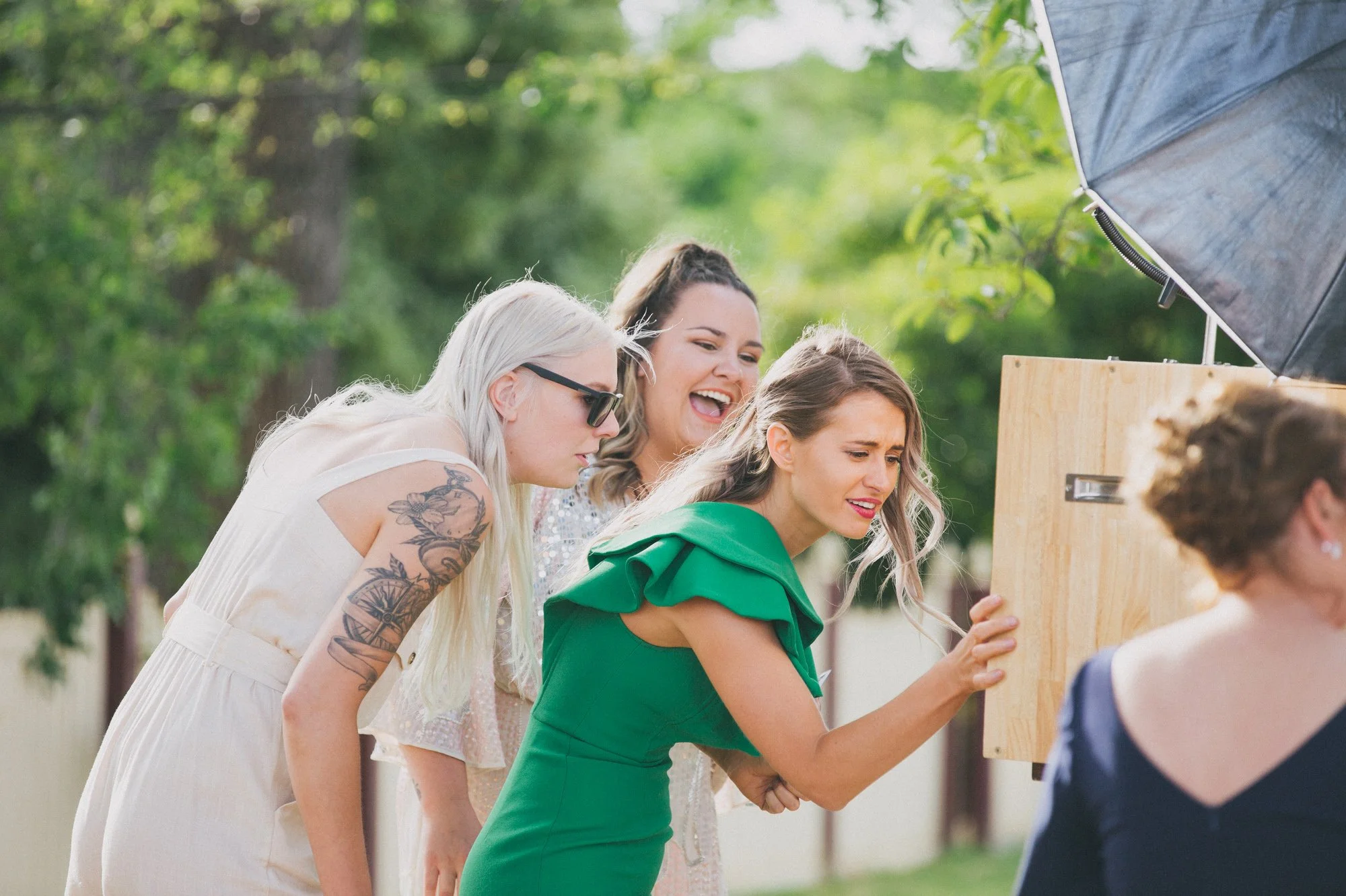 Four women gathered outdoors, looking at a wooden photo booth or display, with trees and greenery in the background. One woman in a green dress appears to be taking a photo or inspecting the display, while the others look on.