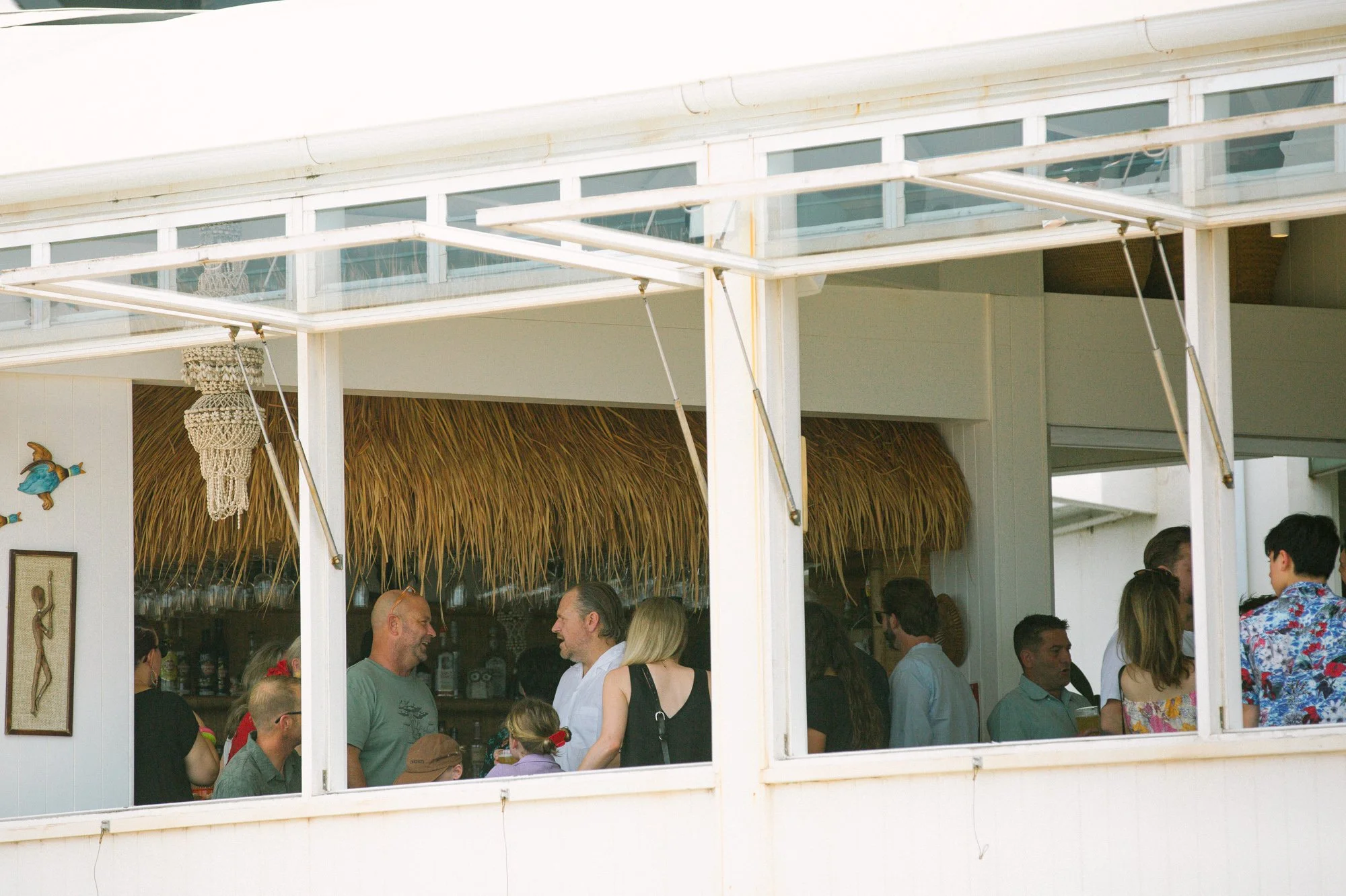 People gathered inside a beachside restaurant or bar with a thatched roof, white walls, and nautical decor, including framed art and hanging ornaments.