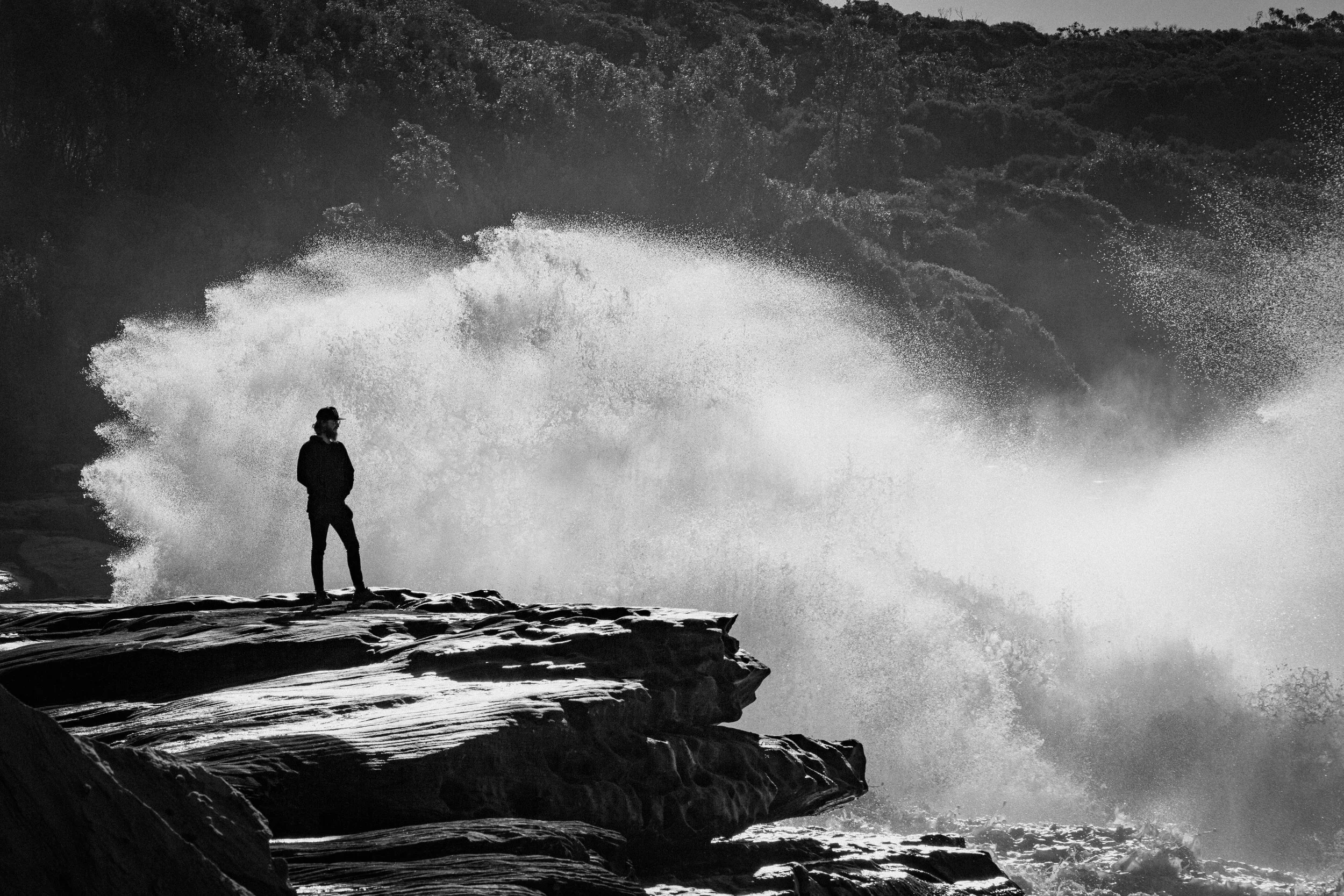 A silhouette of a person standing on rocks near the ocean as large waves crash behind them, in black and white.