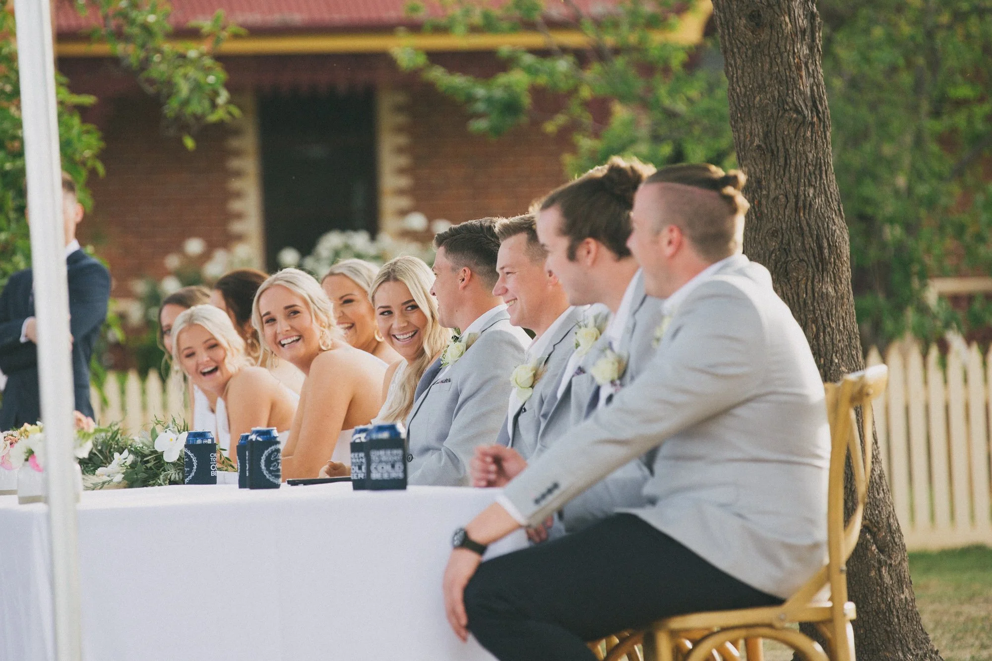 A group of smiling people, including men and women, sitting at a long table outdoors during a wedding ceremony, with some wearing suits and dresses, with flowers and canned drinks on the table.
