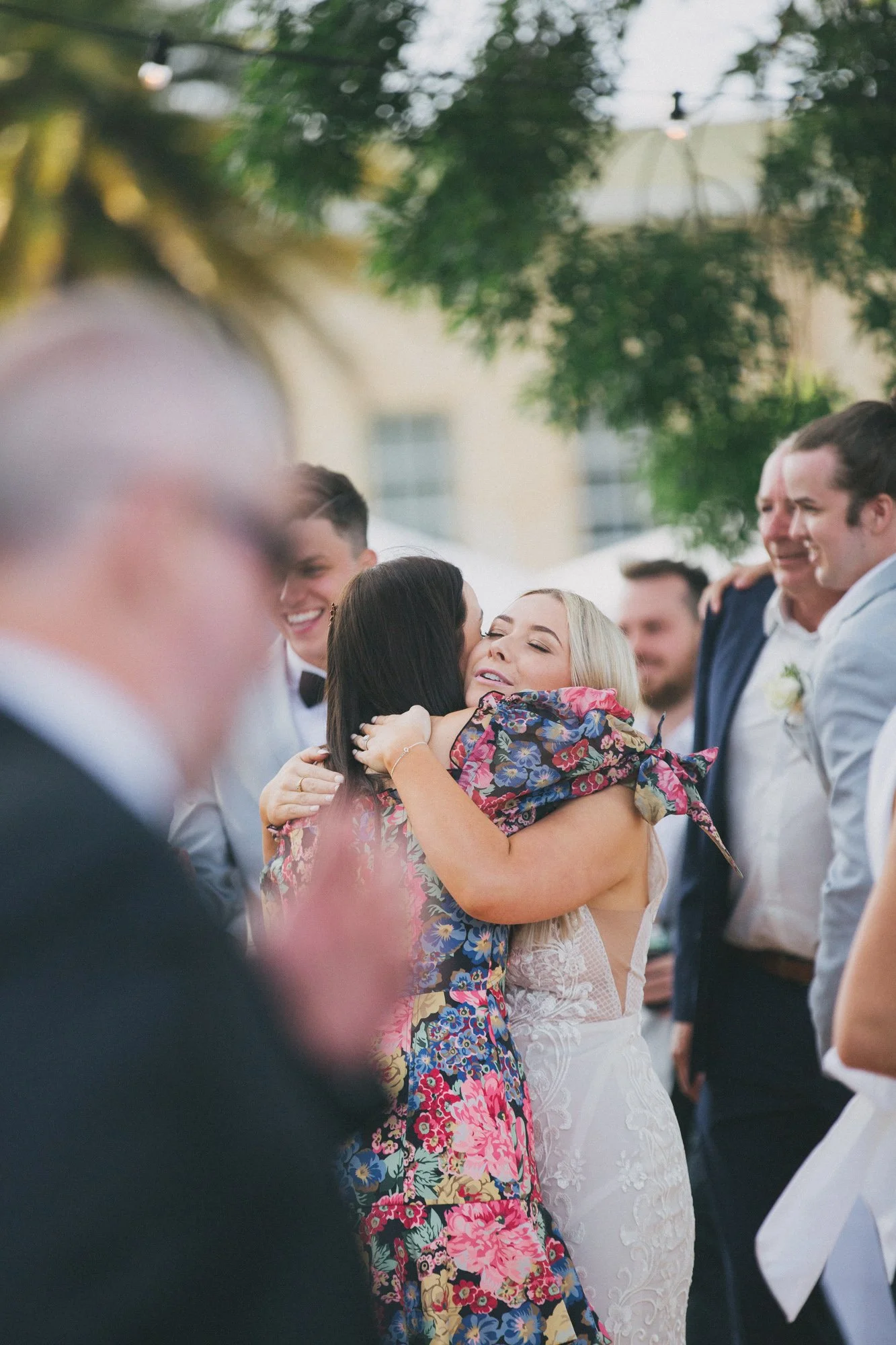 Two women hugging at a wedding, surrounded by friends in formal attire. One woman is wearing a white lace dress, and the other is in a floral dress.