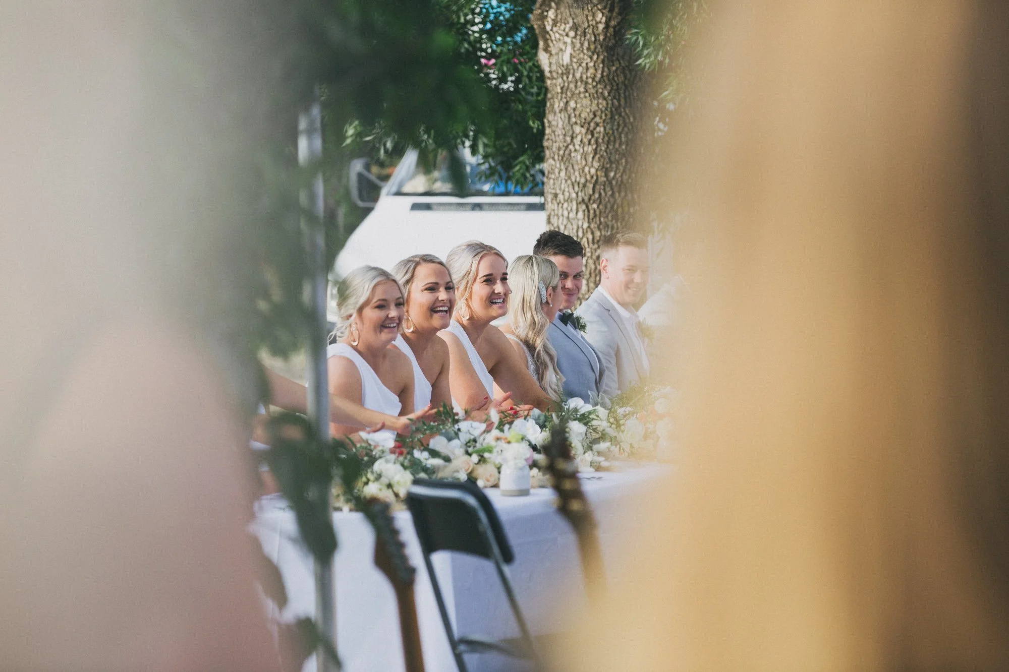 A wedding reception table outdoors with six smiling people, mostly women, dressed in white and light-colored formal attire, seated at a decorated table with flowers, with a large tree and a white vehicle in the background.