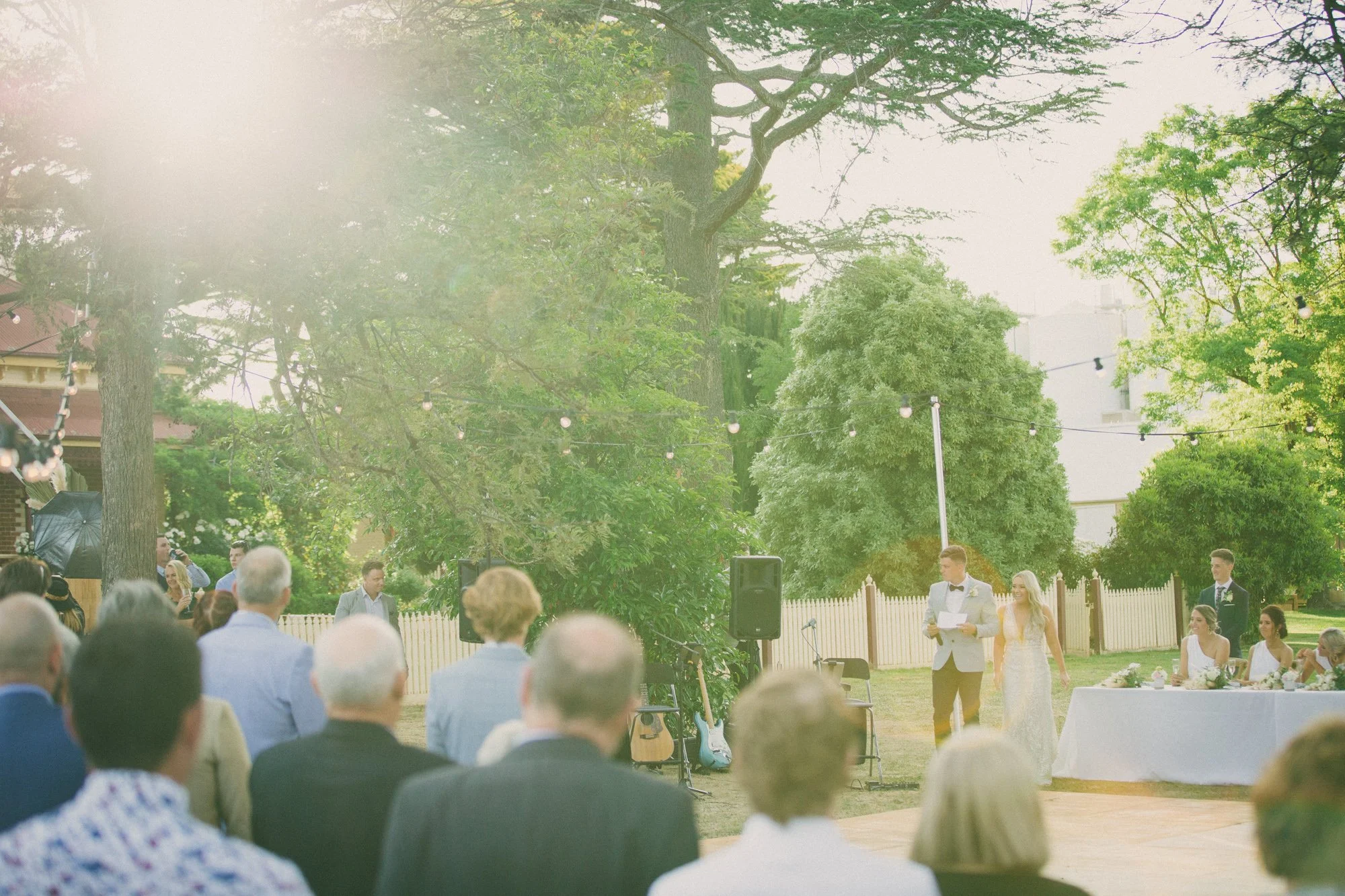 Outdoor wedding ceremony with guests seated and a bride and groom standing at the altar, surrounded by greenery and string lights.