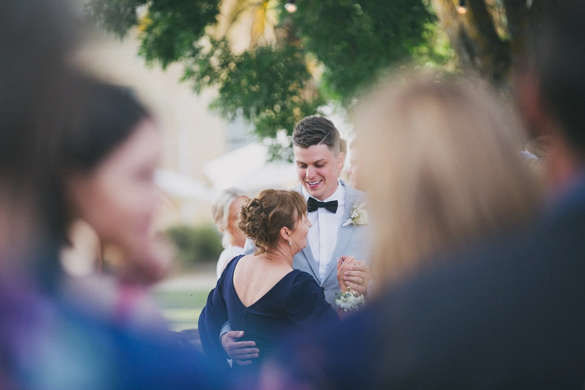 A young man in a tuxedo smiling and dancing with an older woman at an outdoor wedding reception, surrounded by guests.