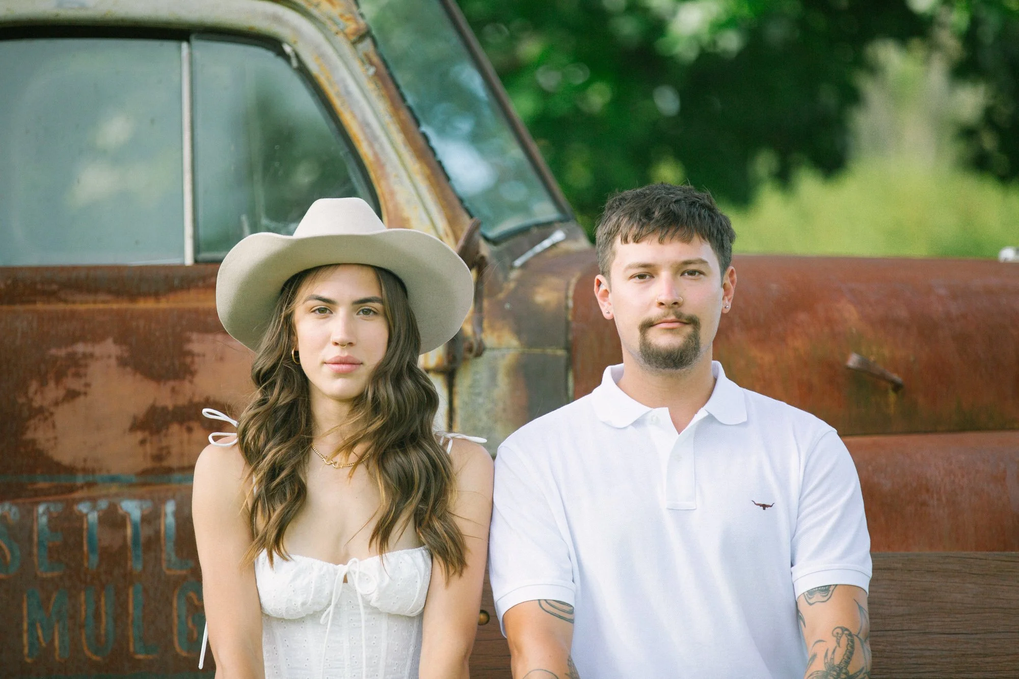 A woman and a man sitting on a wooden bench outdoors, with a rusted old truck behind them, surrounded by green trees.