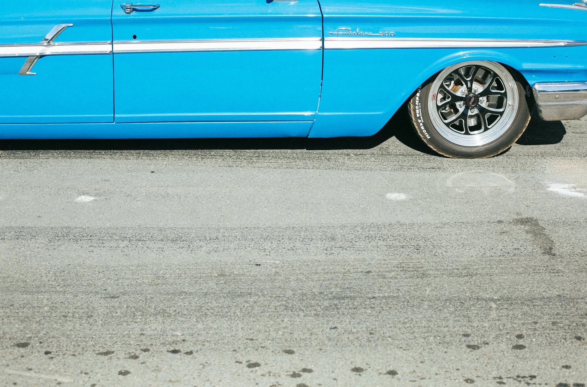 Close-up of the side of a vintage blue car with a 'Fairlane 500' badge, black alloy wheels, and low profile tires, parked on a concrete surface.