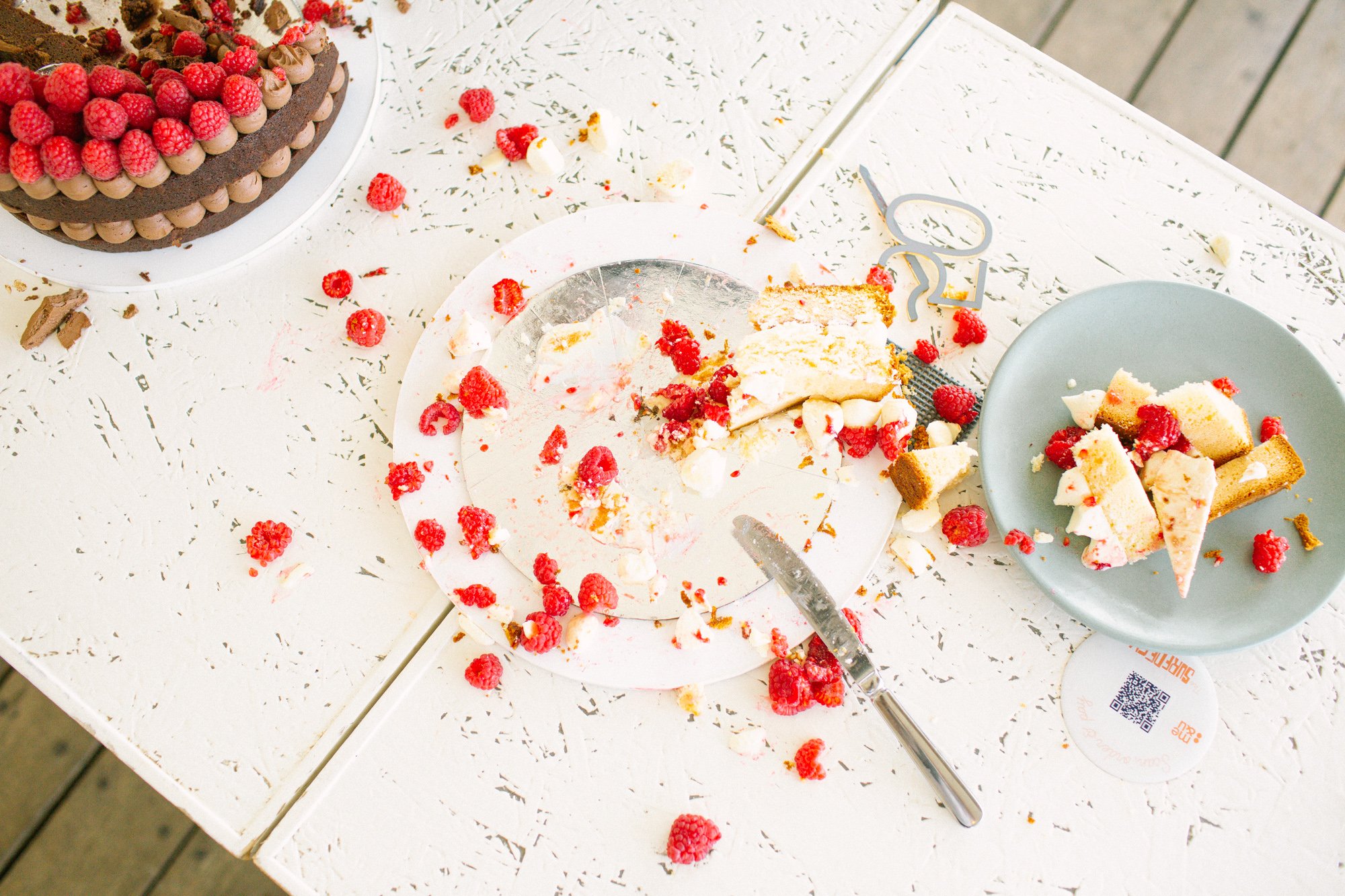 A white table with a toppled birthday cake with raspberries, a plate of cake pieces with raspberries, and a cake cutter, all covered in cake crumbs and raspberry pieces.