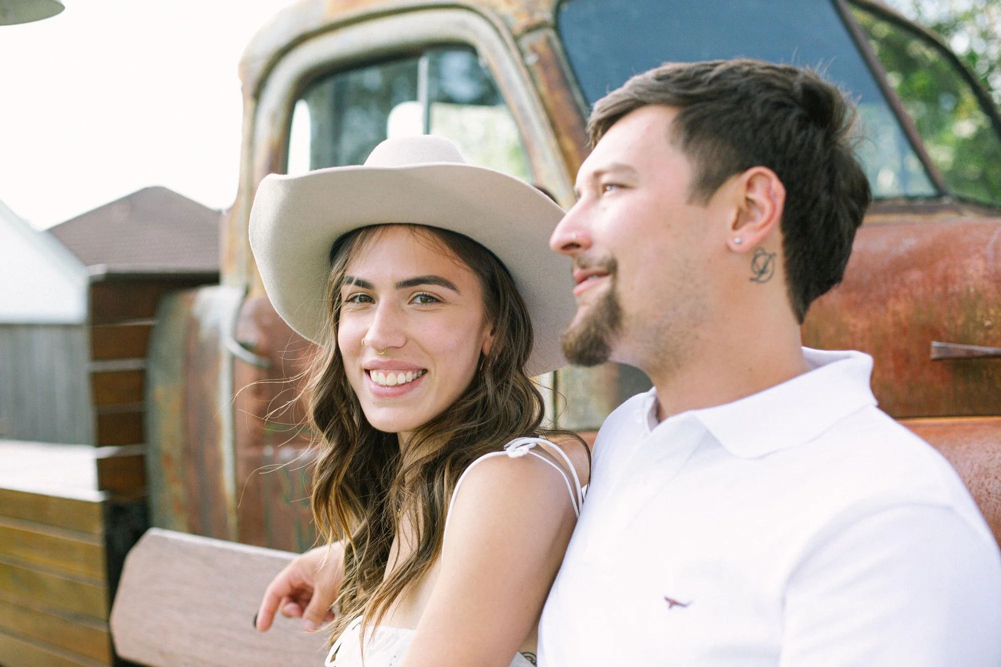 A young woman with long brown hair, wearing a large white hat, and a young man with short dark hair, facial hair, and a tattoo behind his ear, sitting close together outdoors in front of a weathered rust-colored truck, smiling at each other.