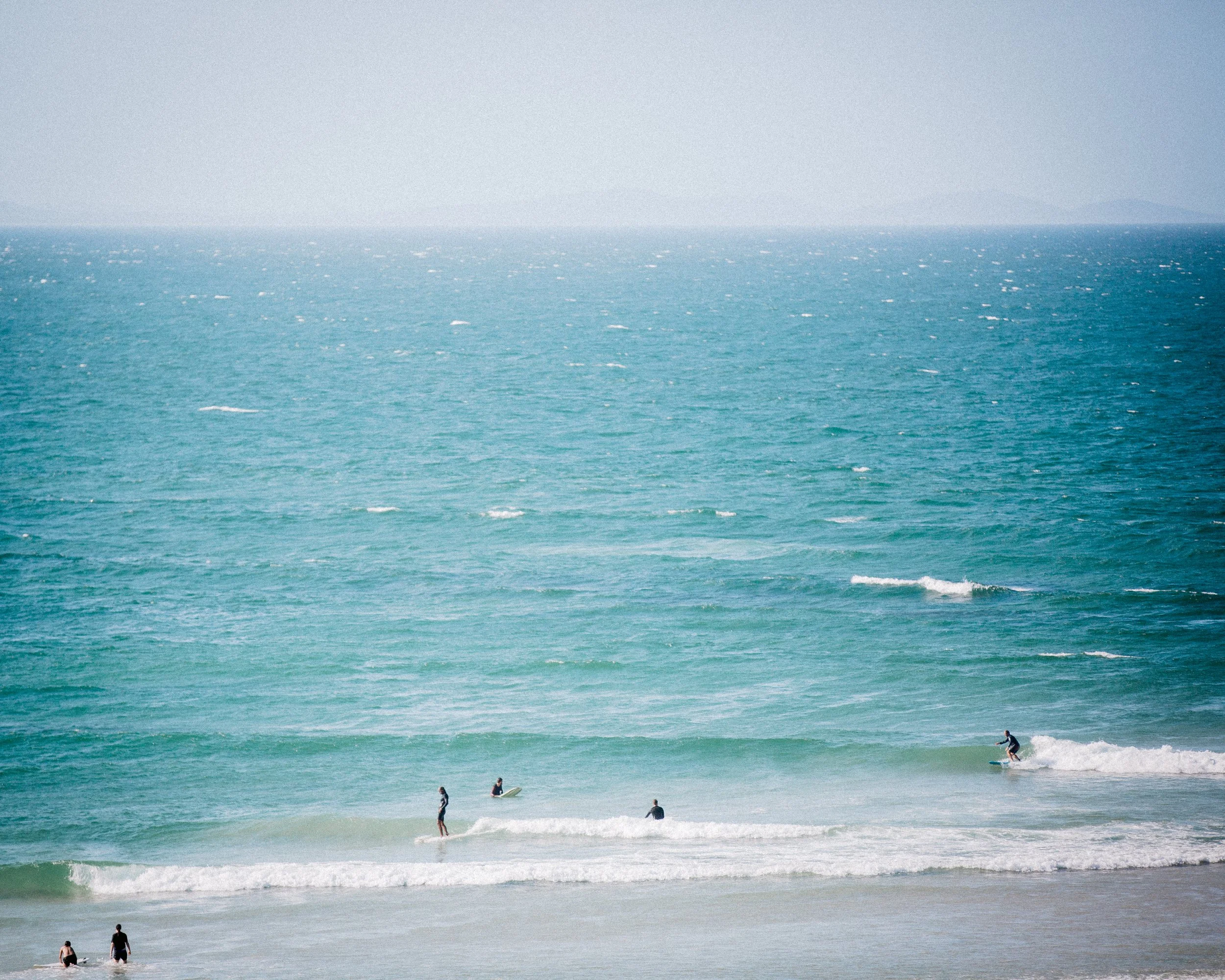 People enjoying the ocean, some surfing and others wading in the shallow water, with the expansive blue sea and distant horizon.