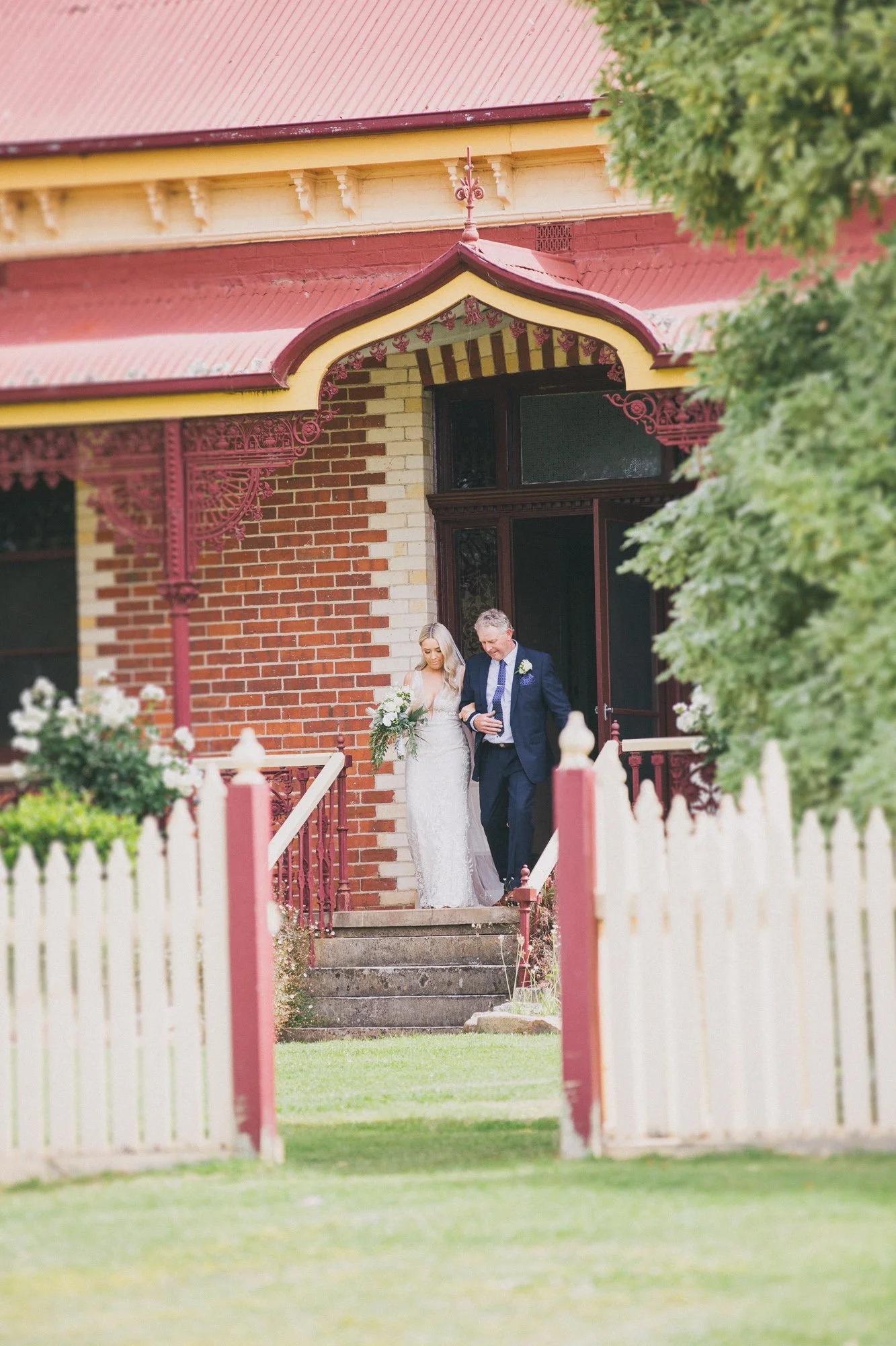 A bride in a white wedding dress holding a bouquet walking down the stairs with an older man in a dark suit outside a red brick house with decorative ironwork and a picket fence.