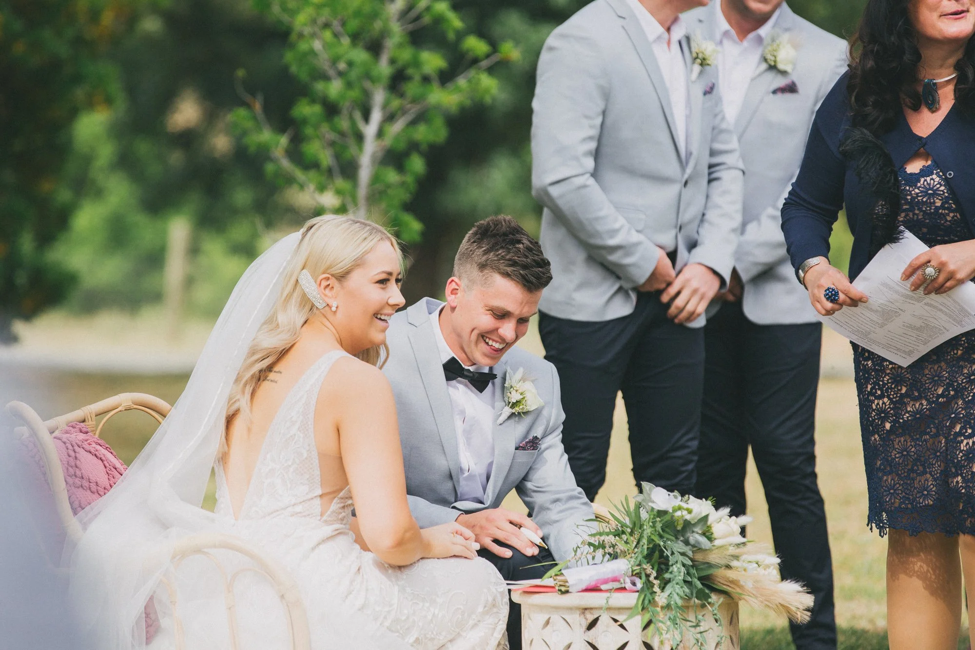 A bride and groom sitting outdoors during their wedding ceremony, smiling and holding hands, with officiant and groomsmen standing nearby.