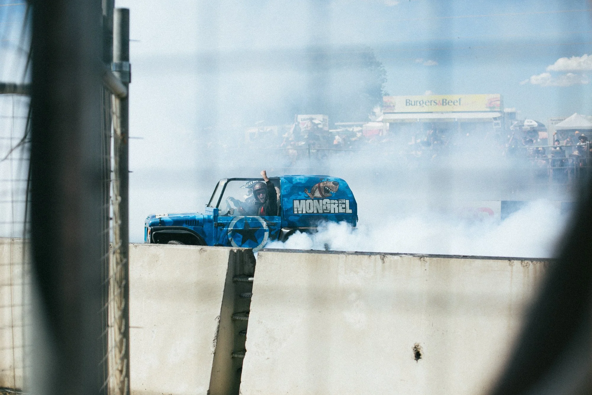 A blue monster truck with the name 'MONGREL' and a cartoon dog graphic on the side, performing a burnout on a race track, producing thick white smoke.