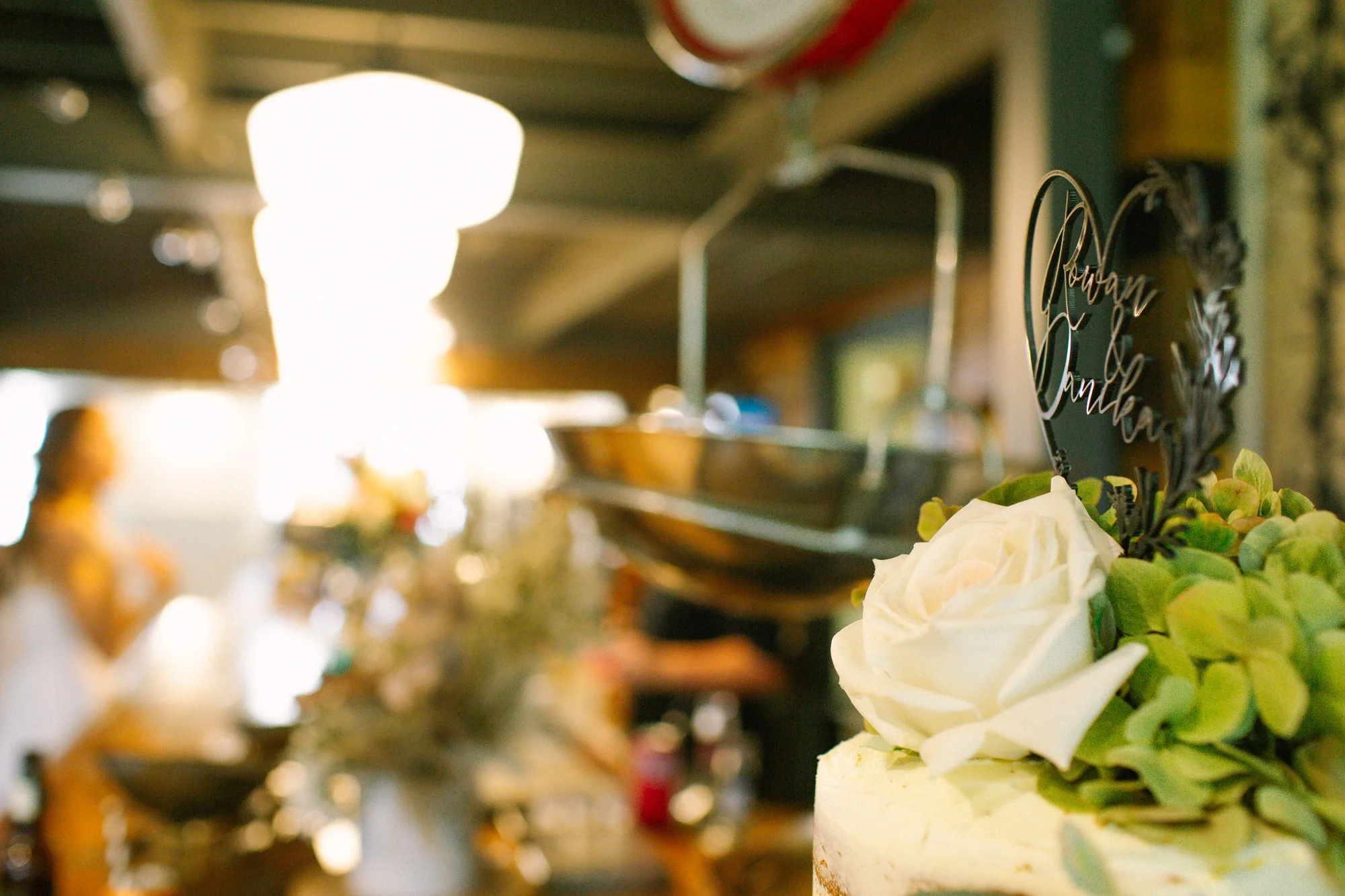 Close-up of a wedding cake with white roses and a Mr. and Mrs. sign topper, with a blurry background of a room decorated for a celebration.