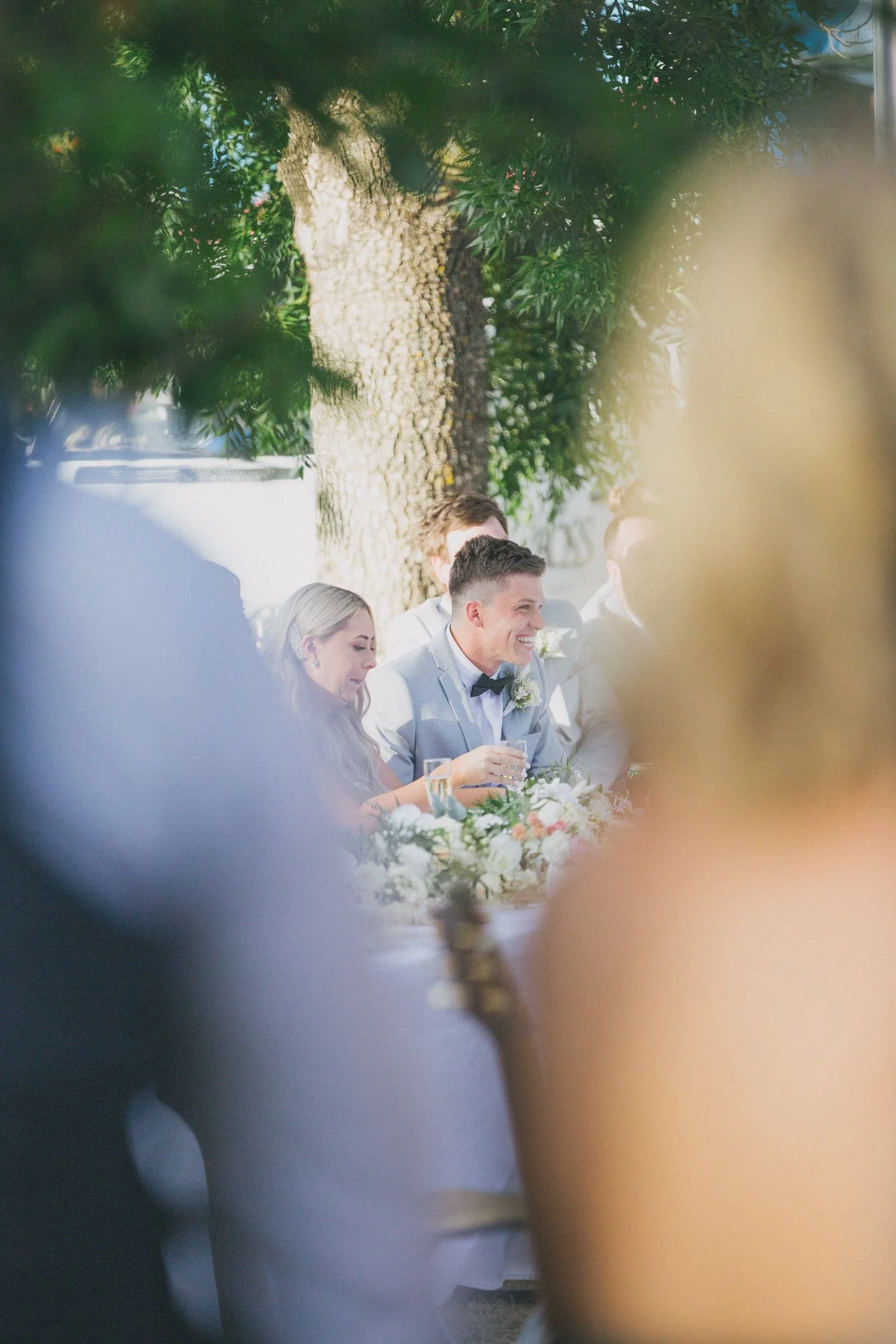 A group of people at a wedding reception, seen through blurred foregrounds, sitting at a table outdoors with a large tree in the background. The focus is on a smiling man in a light gray suit and bow tie, with other guests around him.
