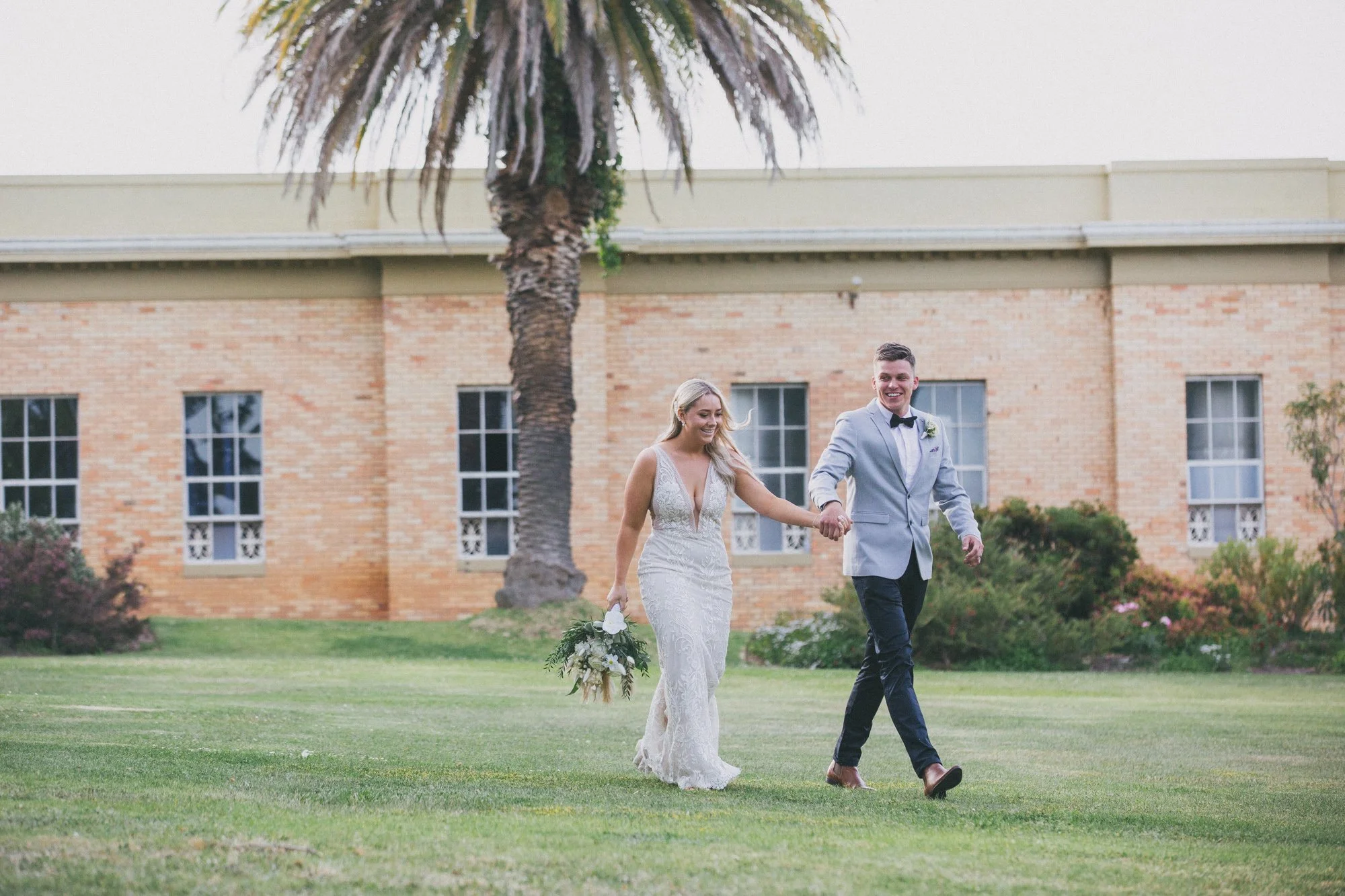 A bride and groom holding hands and walking on a grassy lawn outdoors, with a large palm tree and a brick building in the background. The bride is in a white lace wedding dress holding a bouquet, and the groom is in a gray suit with a black bow tie.