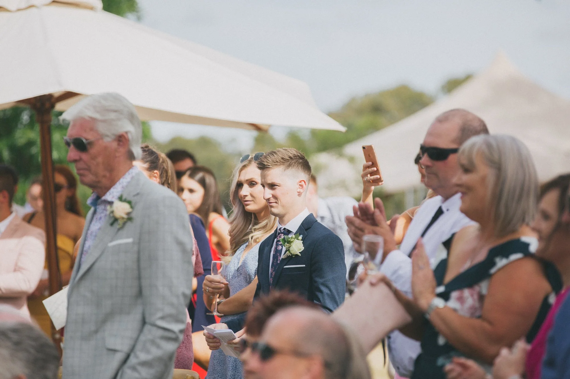 People attending an outdoor wedding ceremony, standing under umbrellas, with some taking photos or holding programs.